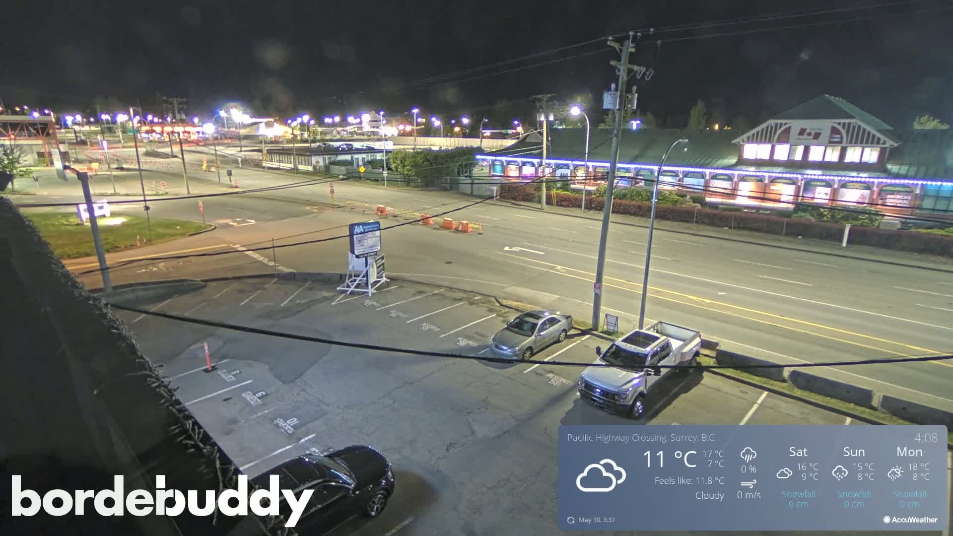 A nighttime, cloudy view of a parking lot and roadway next to a brightly lit building, with several cars visible.