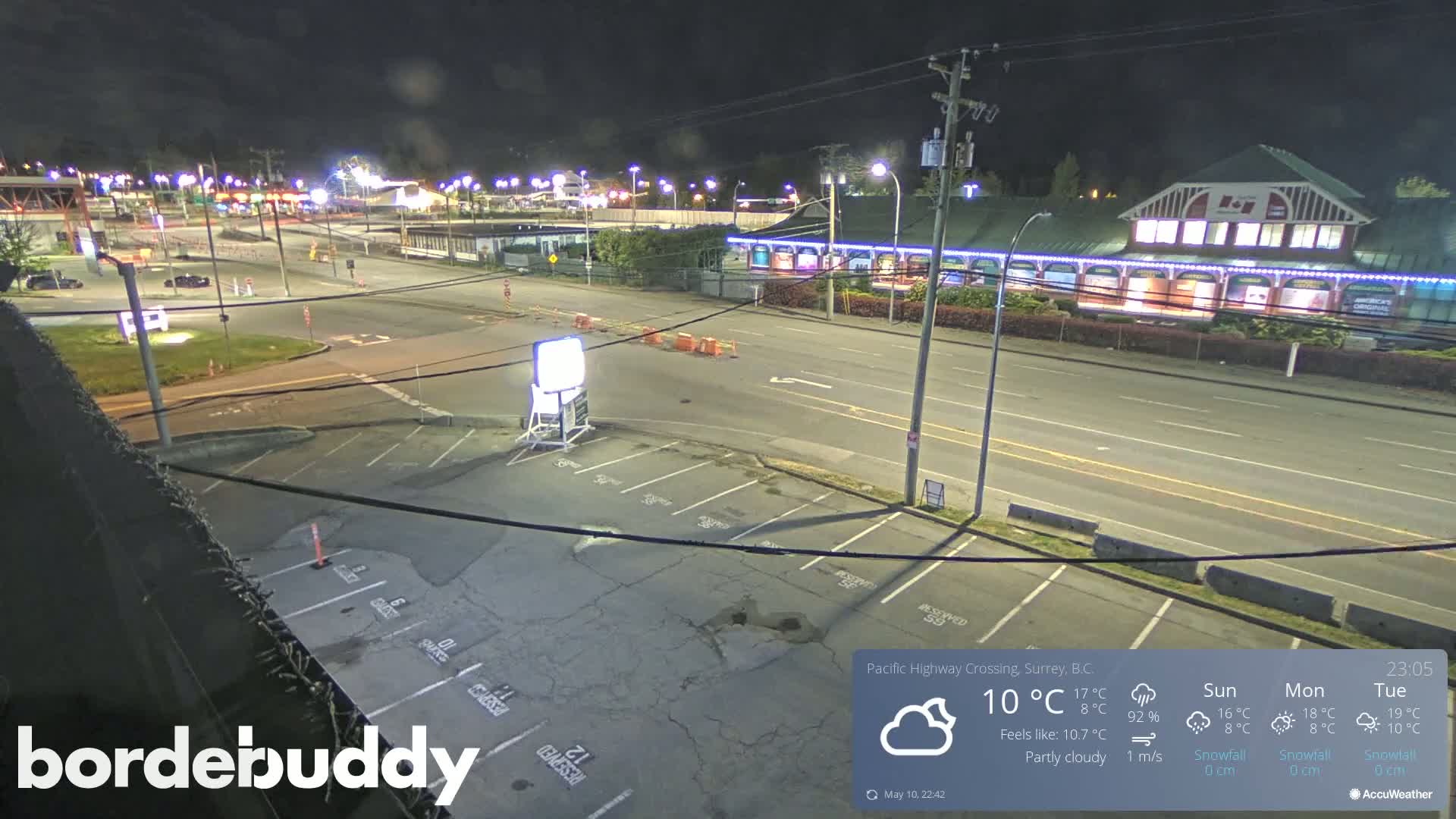 A nighttime, partly cloudy view of a highway crossing with a large building and parking lot, showing some traffic and construction barriers.