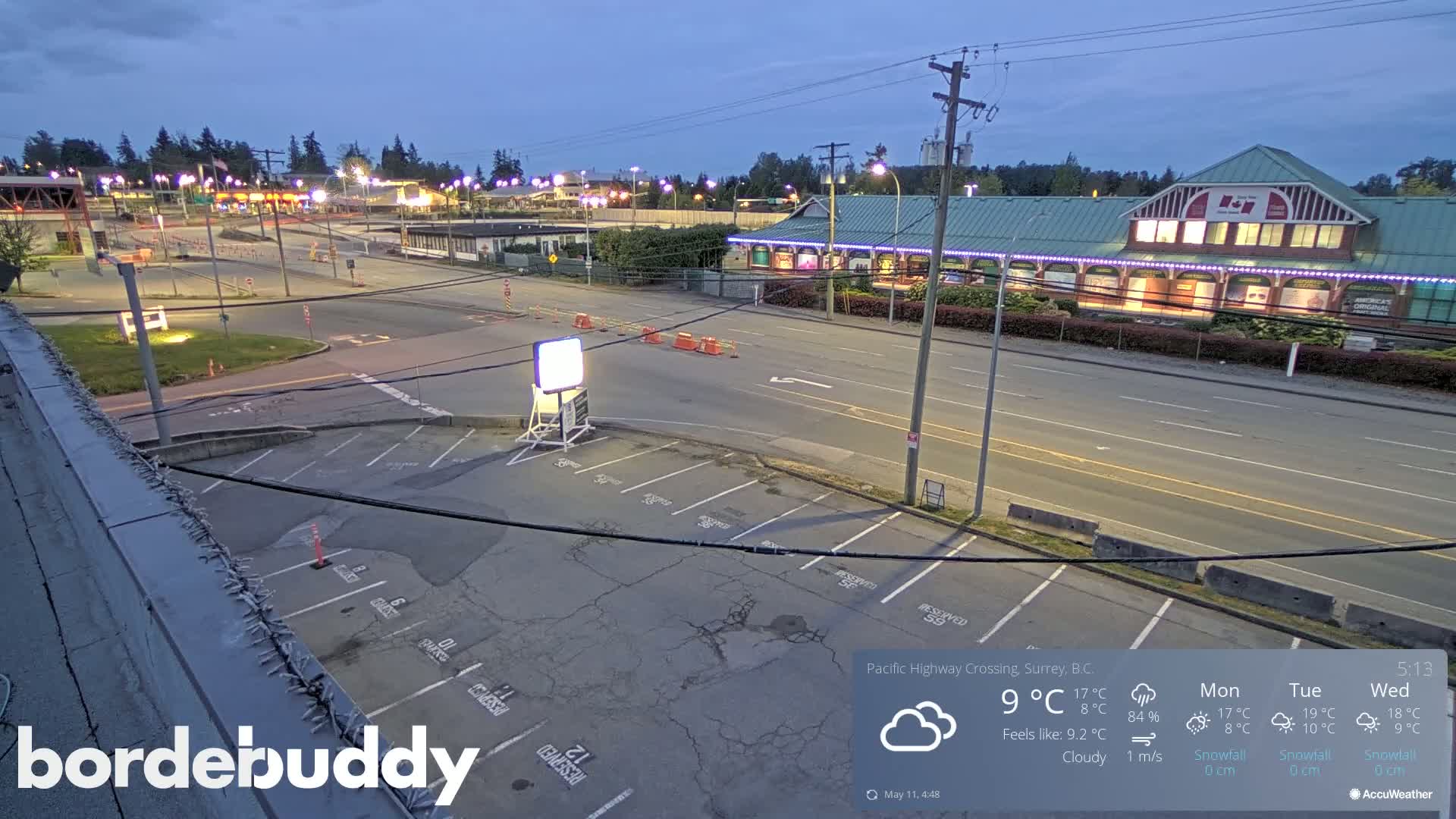 An empty parking lot overlooks a mostly empty street and a building with lights under a cloudy evening sky.