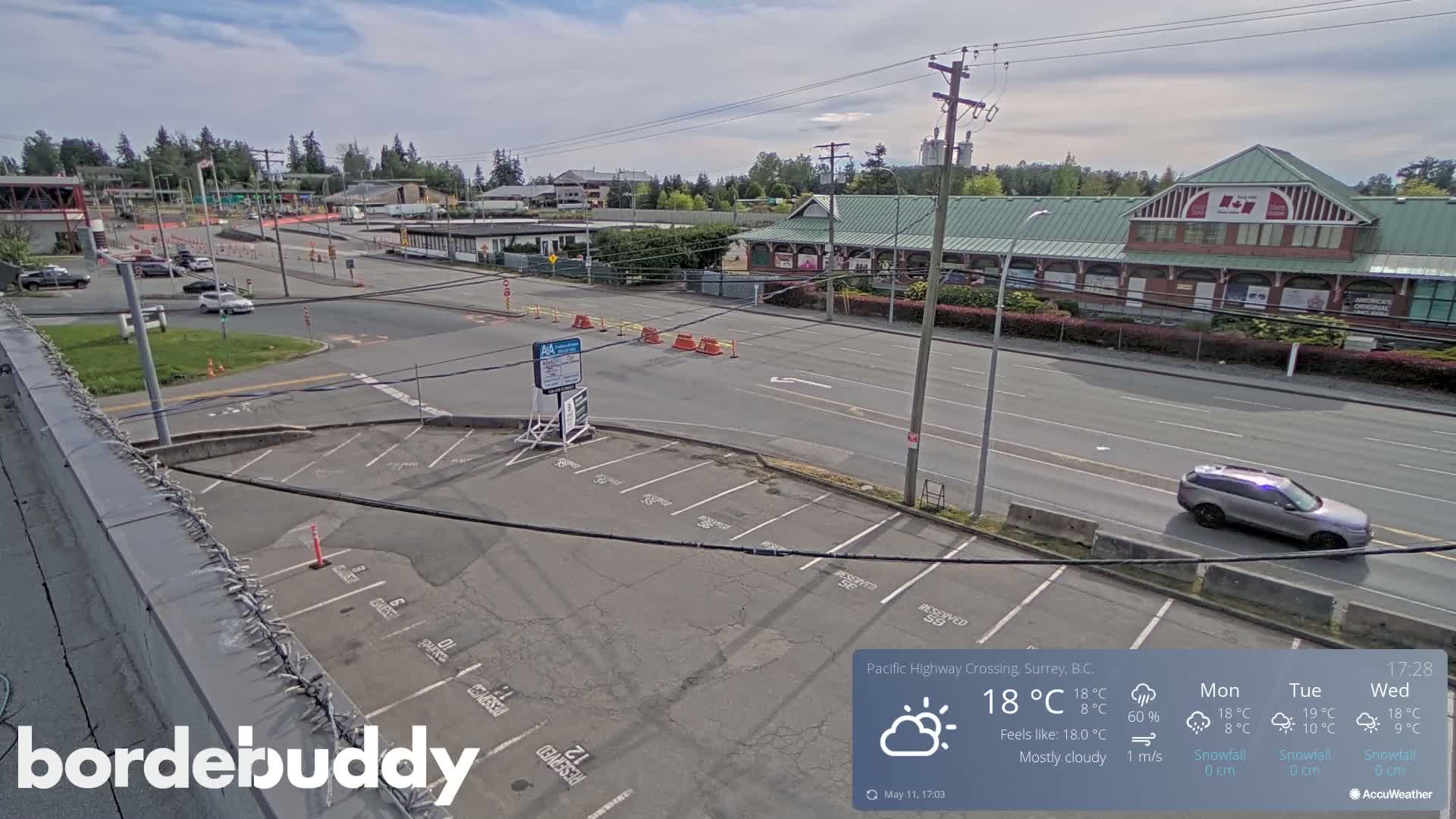A mostly cloudy day shows a parking lot and roadway with several vehicles, buildings, and some construction barriers.