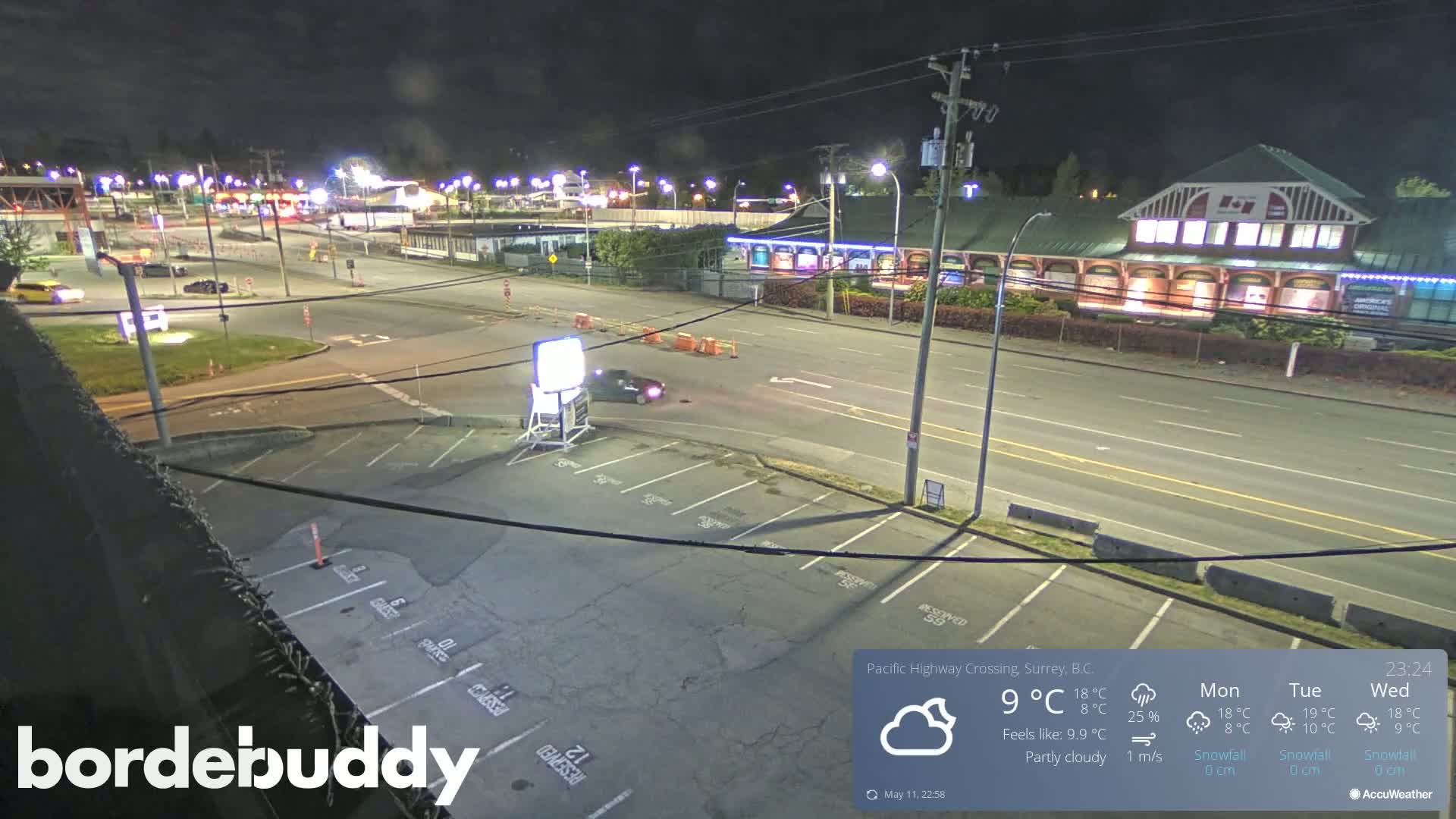 A night-time view of a mostly empty parking lot and street, with a few cars visible, under partly cloudy skies.