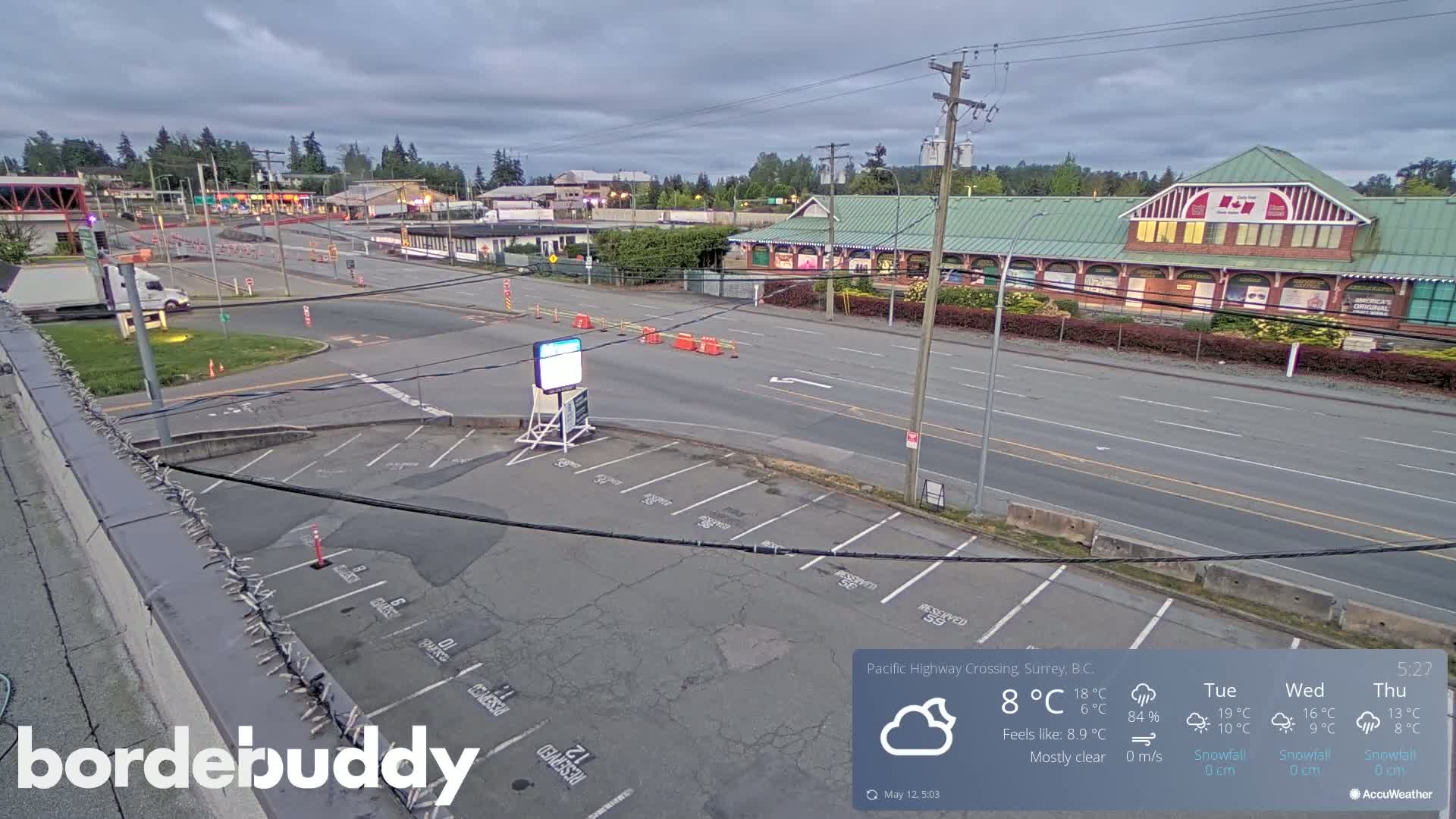 An empty parking lot and road are seen under mostly clear skies, with a few buildings in the background.