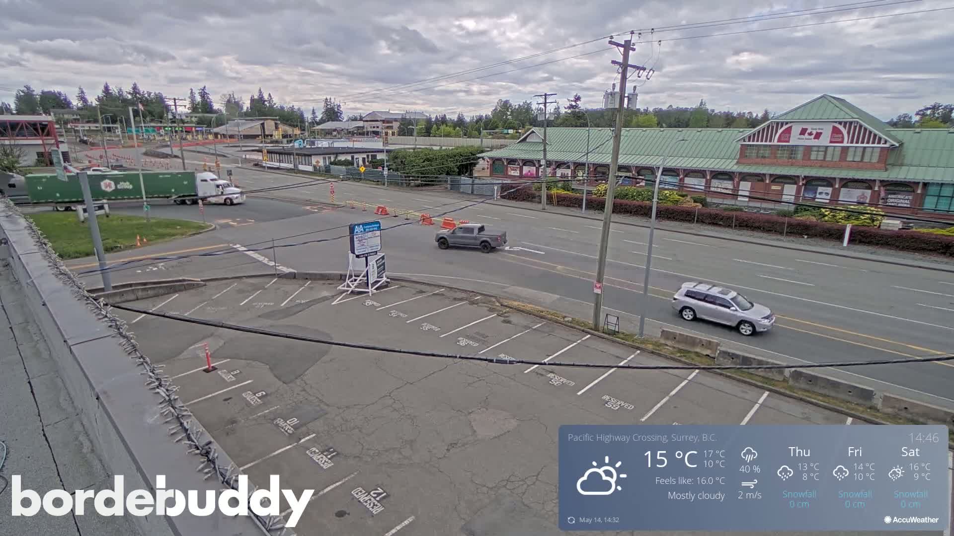 A mostly cloudy day reveals a mostly empty parking lot overlooking a highway intersection with several vehicles, including a semi-truck, and a large brick building.