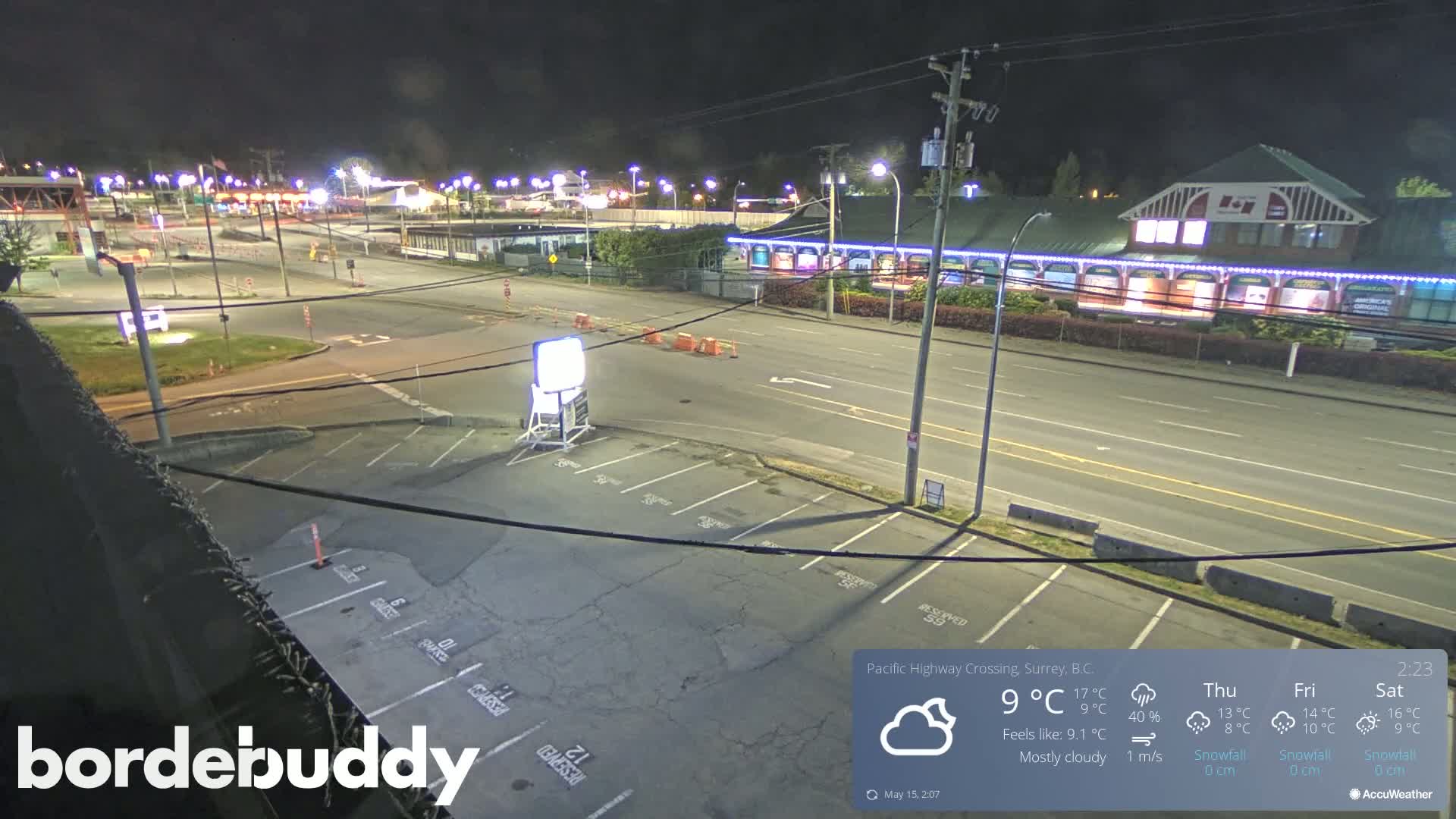 A mostly cloudy night scene shows a mostly empty parking lot next to a multi-lane road with buildings illuminated along the opposite side.