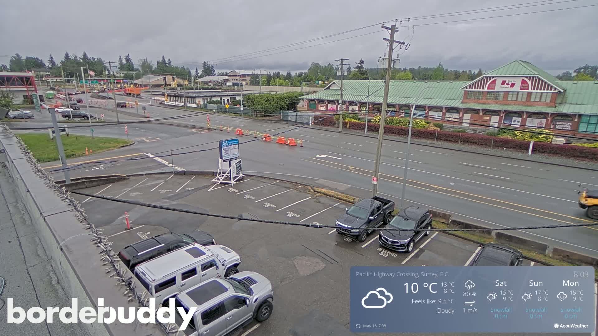 A cloudy day shows a street scene with a parking lot containing several cars, a building with a green roof, and a roadway with some construction.