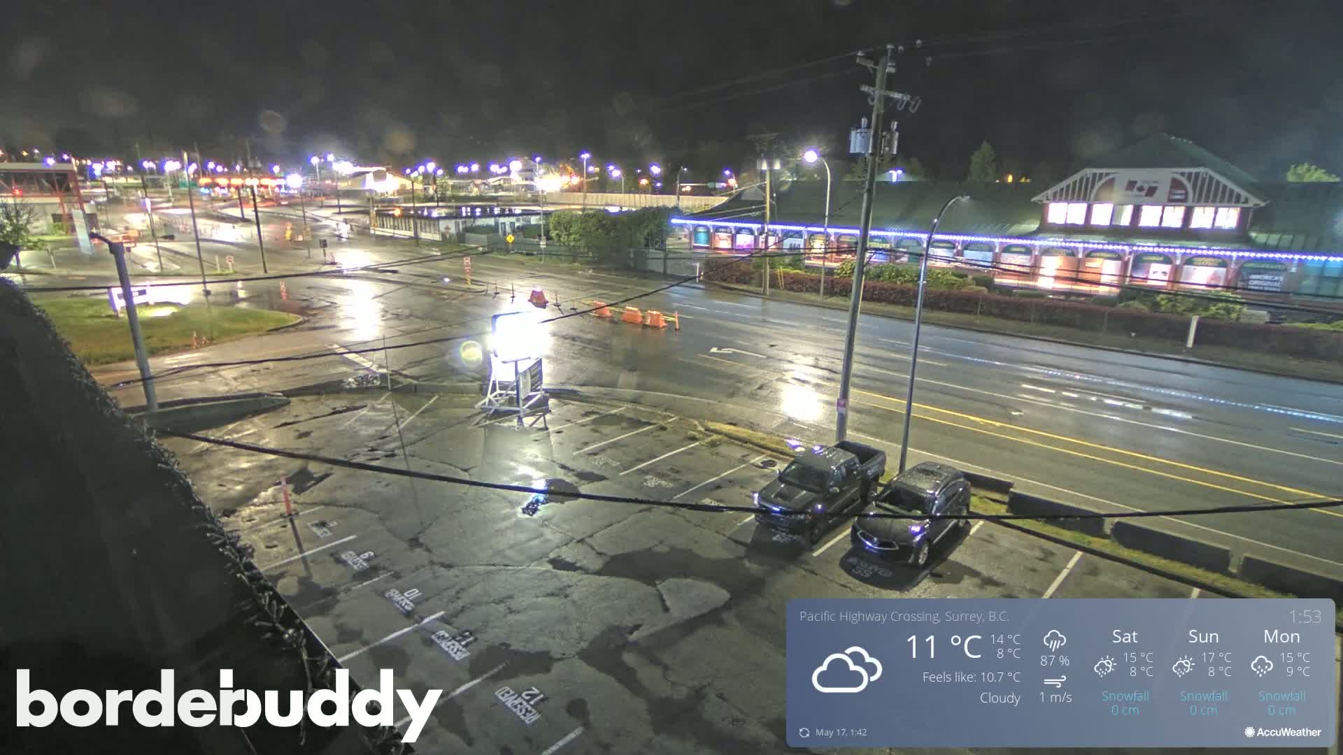A nighttime, rainy view of a parking lot and street, with cars parked and a building with lights visible in the distance.