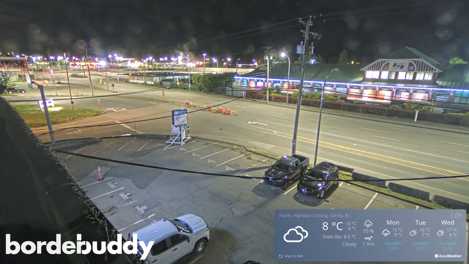 A nighttime, cloudy scene shows a parking lot with several vehicles and a street with buildings and streetlights visible in the distance.