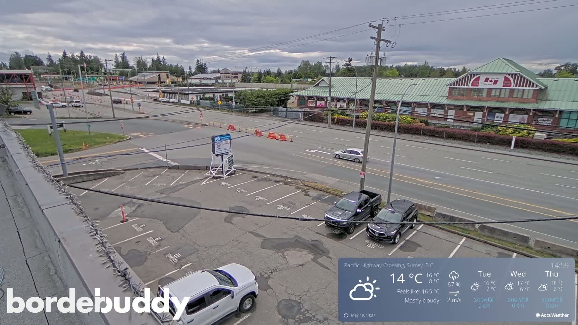 An elevated view of a mostly cloudy parking lot next to a roadway with several vehicles, buildings, and road construction visible in the distance.