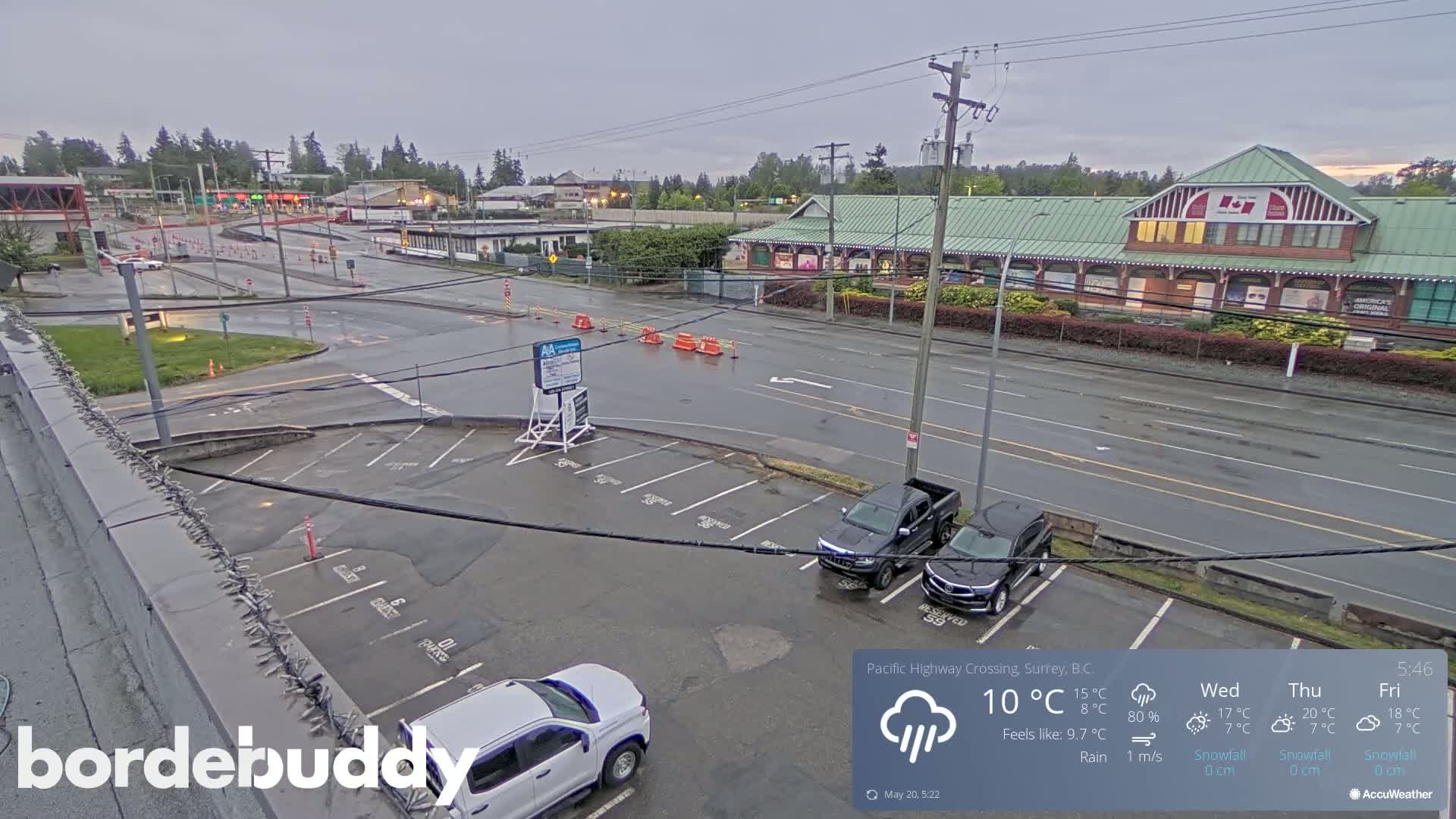 A wet parking lot with several vehicles is shown next to a roadway with light traffic under a cloudy sky.