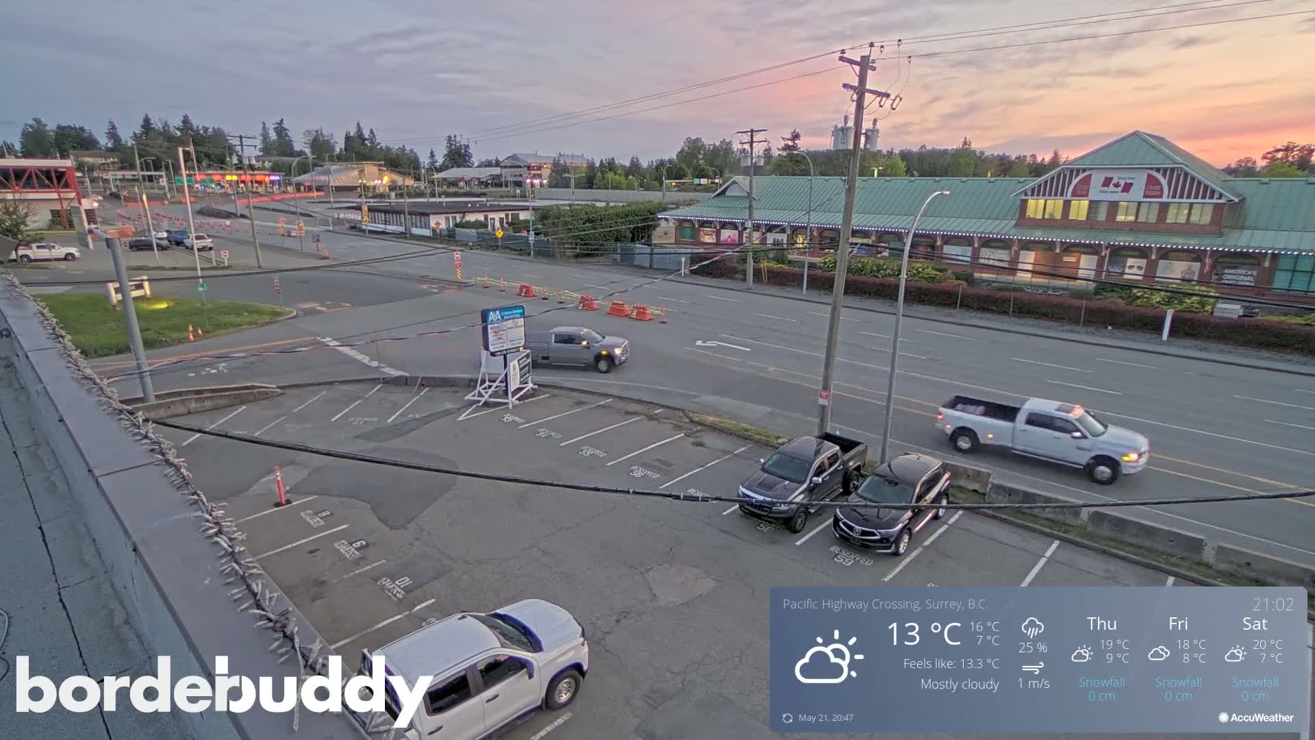 A mostly cloudy twilight scene shows a parking lot with several vehicles, and a roadway with more vehicles, including pickup trucks, near buildings.