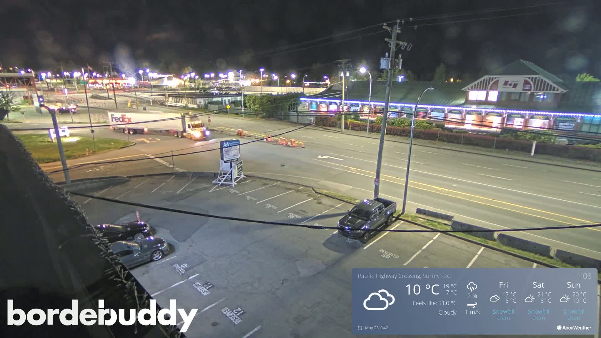 A nighttime, cloudy scene shows a FedEx truck turning onto a road near a parking lot with a few cars, some buildings, and streetlights.