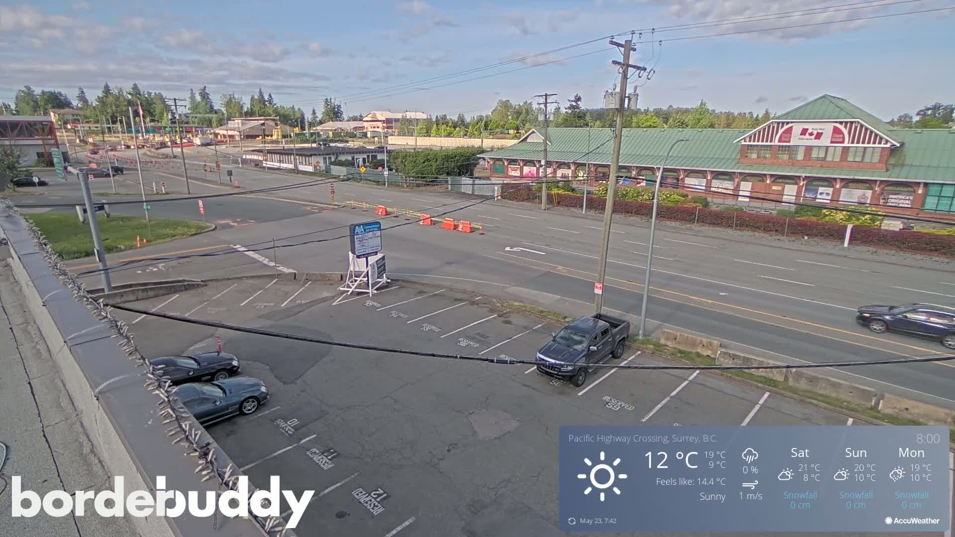 A sunny, overhead view of a roadway intersection with a parking lot, several vehicles, and a large building in the background.