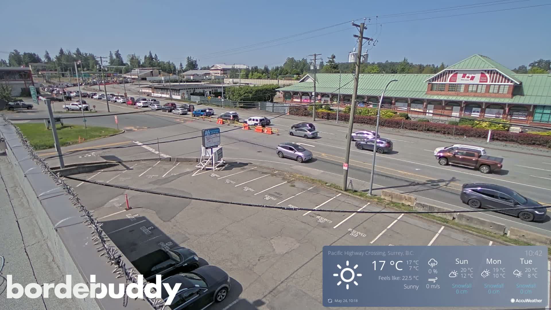 A sunny day shows a parking lot and roadway with several cars, a building with a green roof, and some trees in the background.
