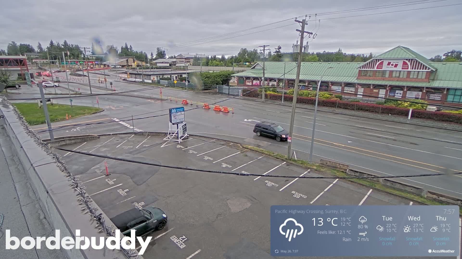 A rain-slicked parking lot and roadway are seen, with a few vehicles present near a building under overcast skies.