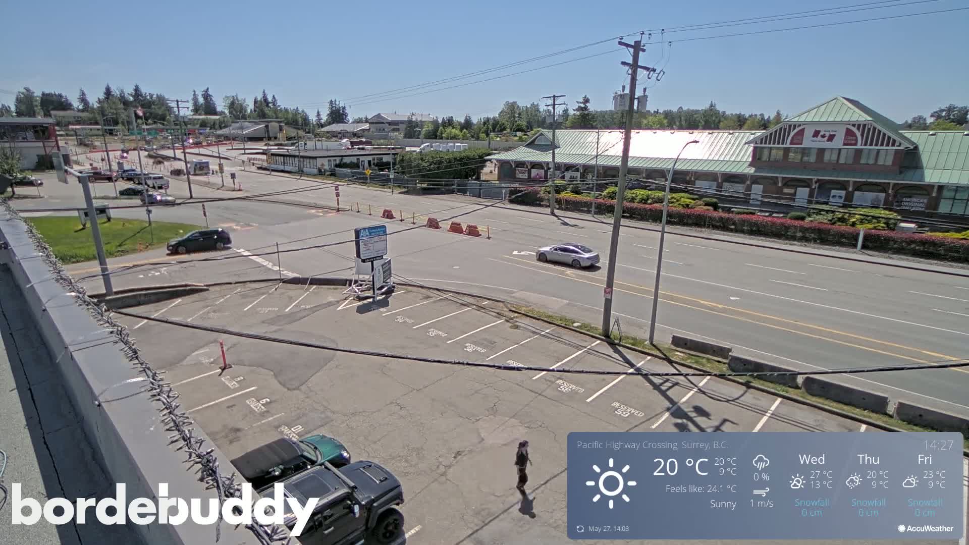 An elevated view of a sunny parking lot and roadway, with several cars visible, next to a large building under a clear blue sky.