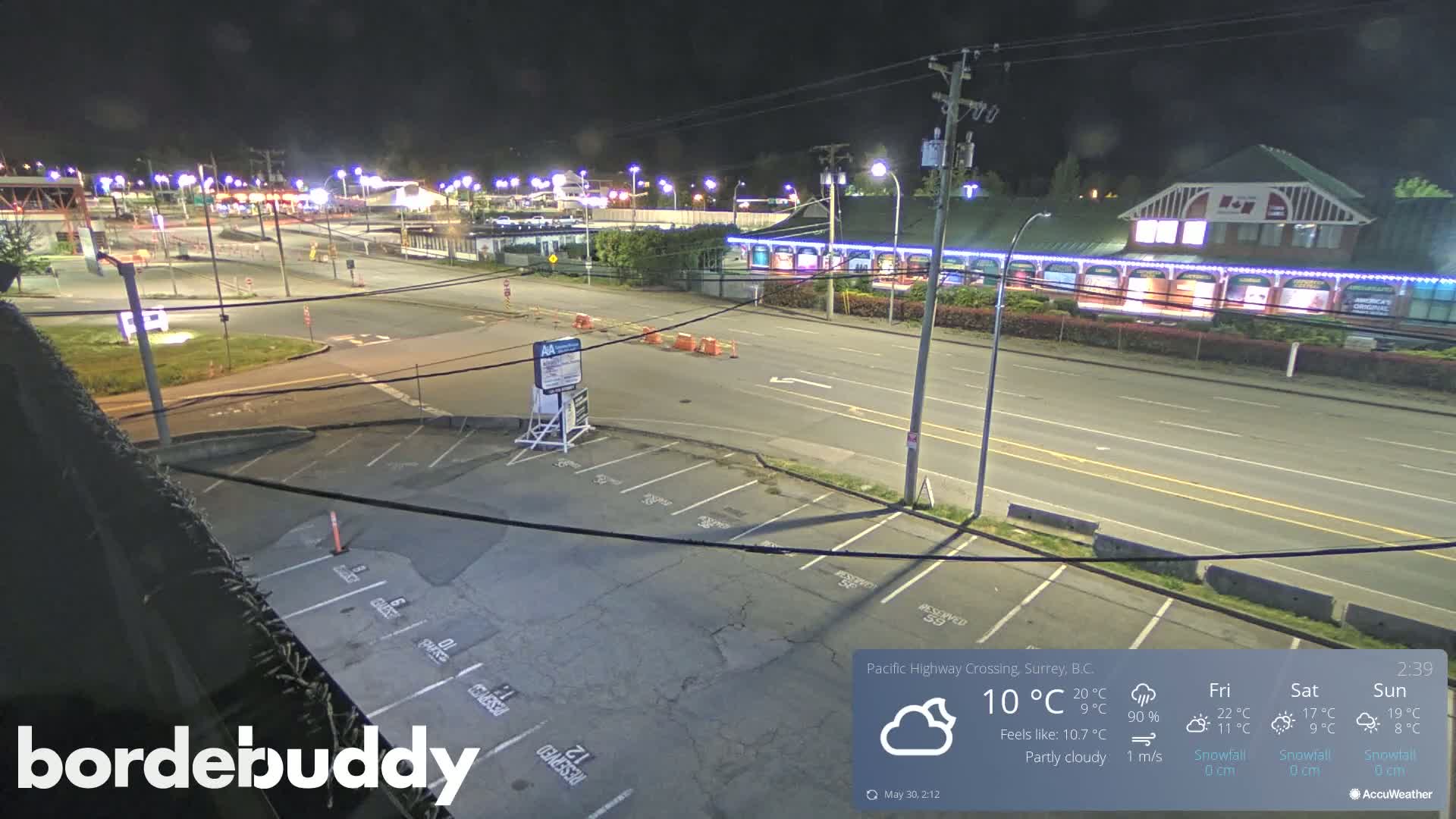 A nighttime, partly cloudy scene shows a mostly empty parking lot next to a roadway with buildings and streetlights visible in the background.