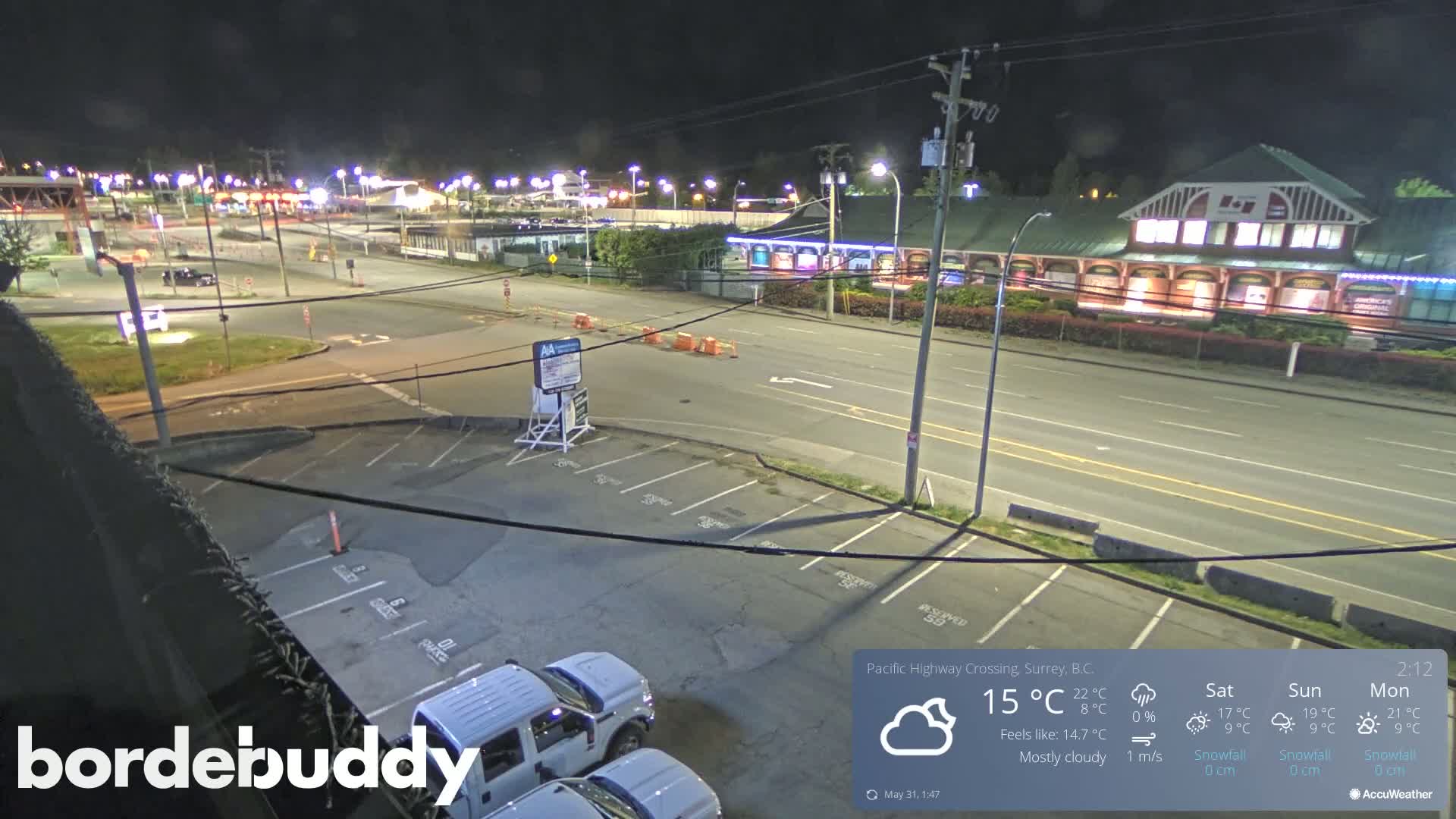A mostly cloudy nighttime scene shows a mostly empty parking lot next to a multi-lane road with buildings and streetlights in the background.