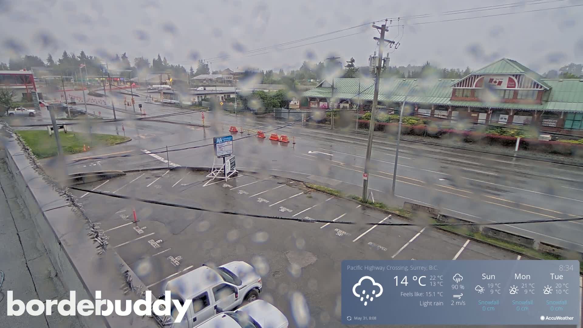 A wet parking lot and roadway are seen from an elevated vantage point during a light rain, with several buildings and vehicles visible in the background.