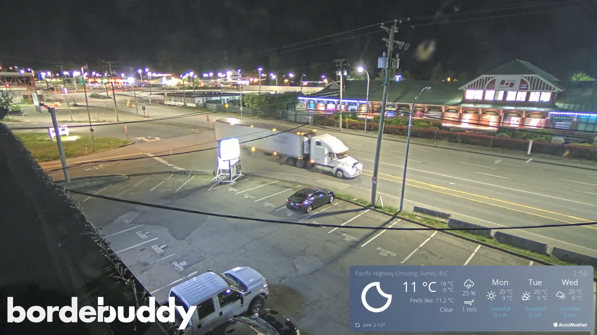 A nighttime, clear-weather view of a highway intersection with a truck and several cars in a parking lot, and buildings in the background.