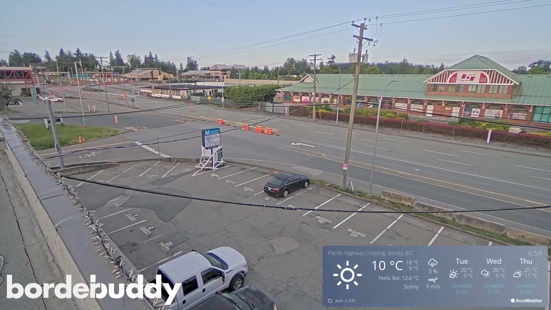 A sunny parking lot and roadway are seen with a few vehicles, a building with a green roof, and some orange traffic barriers in the distance.
