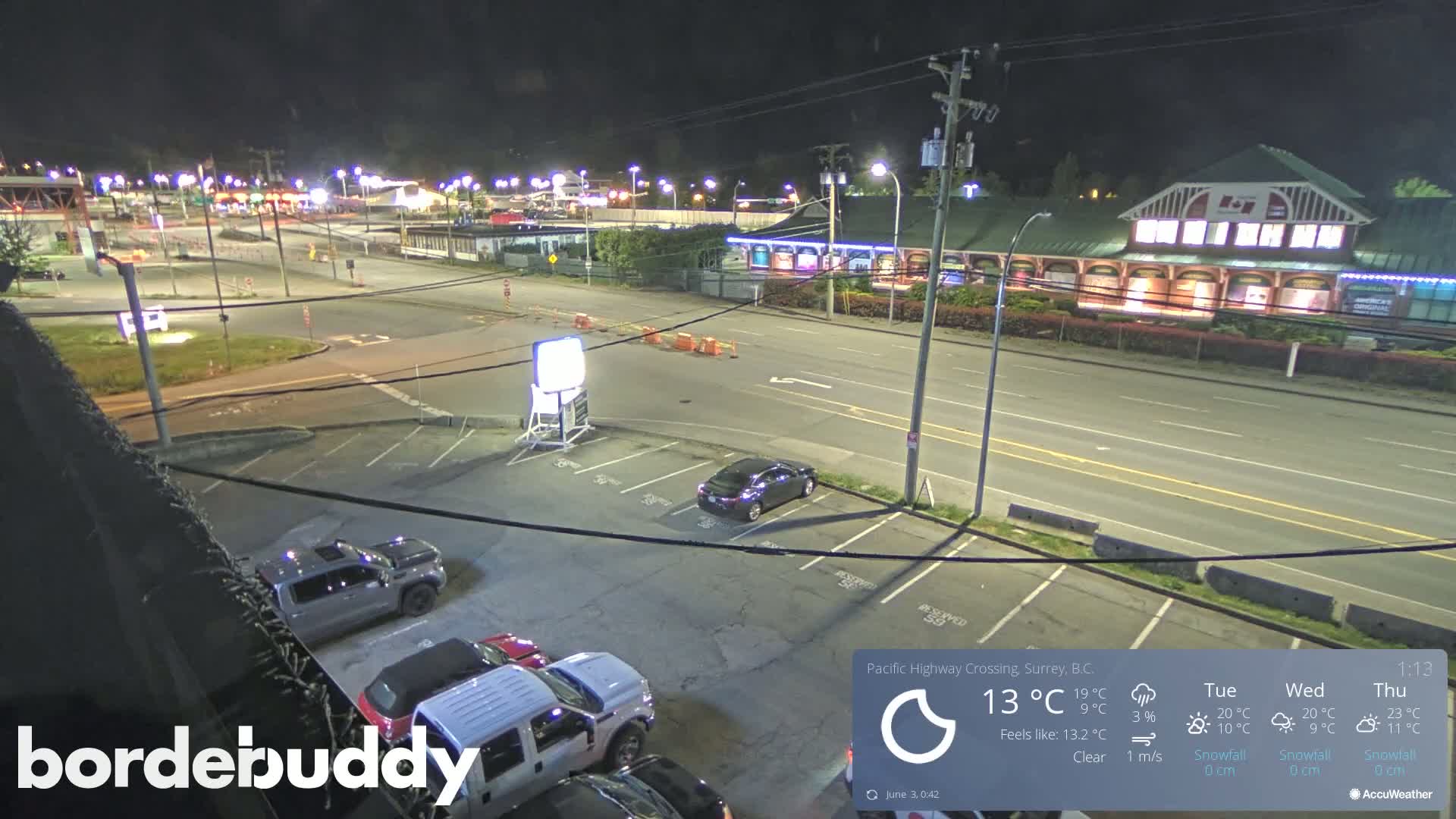 A nighttime, clear view of a parking lot and roadway, with several vehicles present, next to a brightly lit building.