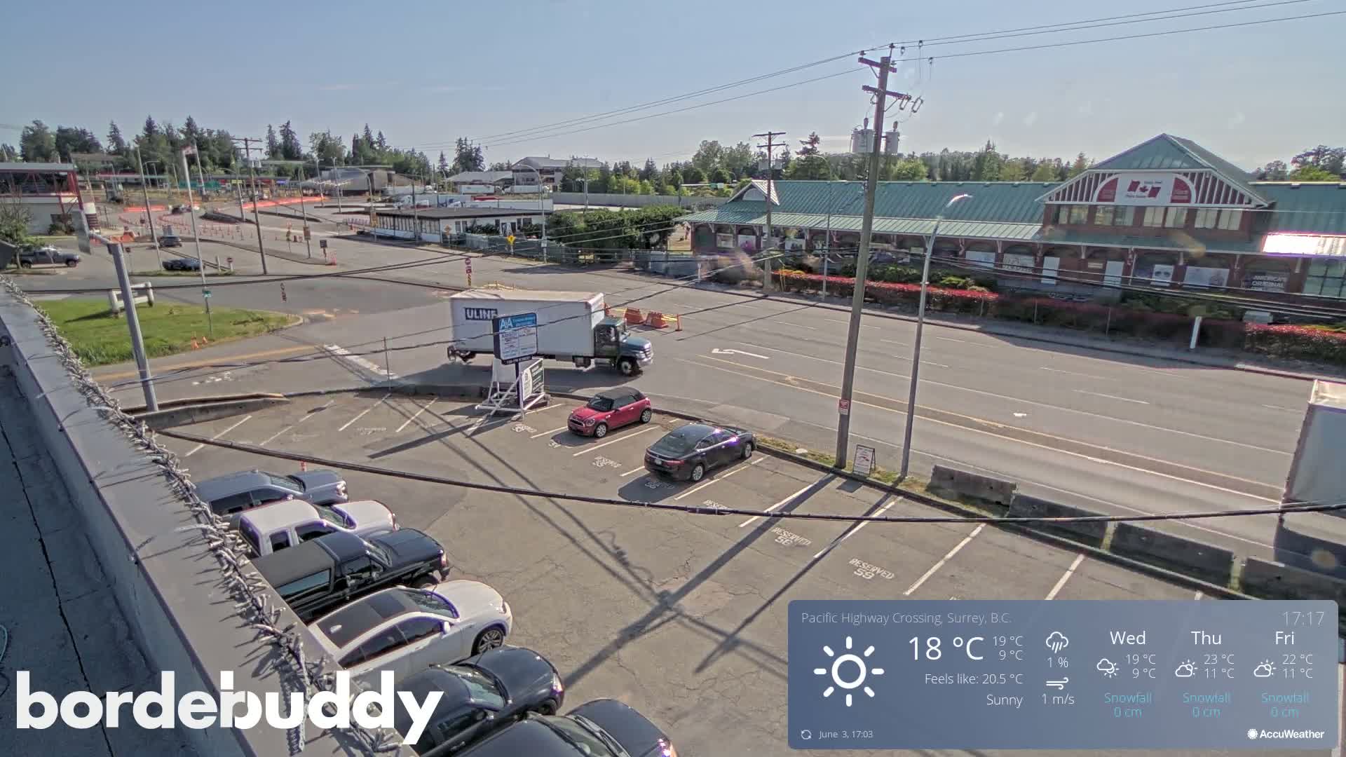A sunny parking lot with several cars and a street scene beyond, including a large building and some trucks.