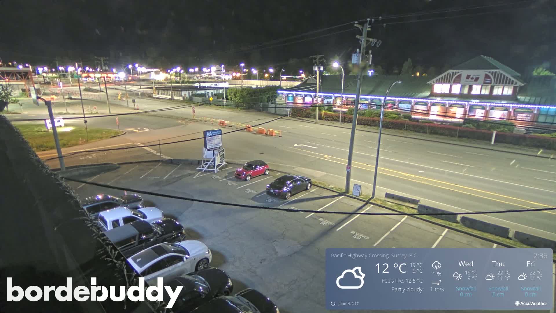 A nighttime, partly cloudy scene shows a parking lot with several cars, a roadway with some traffic, and a lit-up building in the background.
