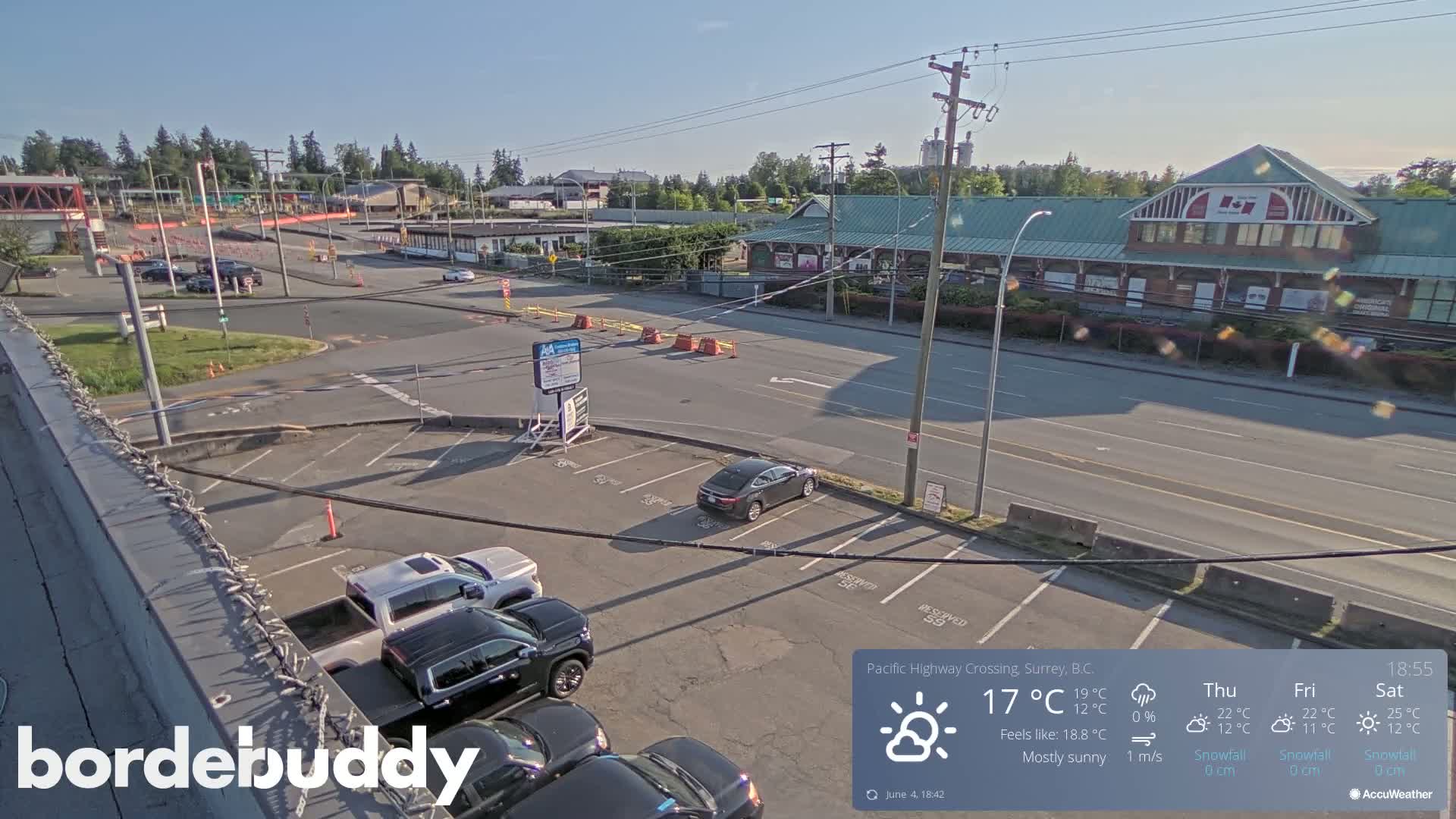 A mostly sunny overhead view of a parking lot, roadway, and buildings, with several vehicles in the parking lot.