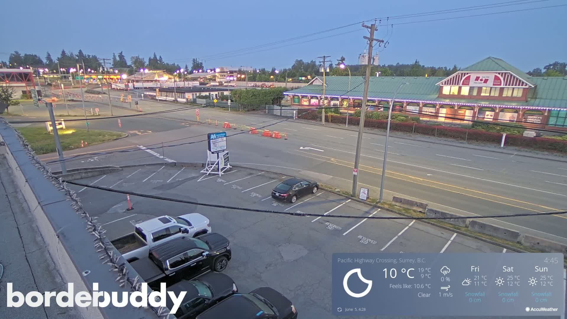 An empty parking lot and roadway are visible at dusk under clear skies, with several vehicles parked and a building with string lights in the background.