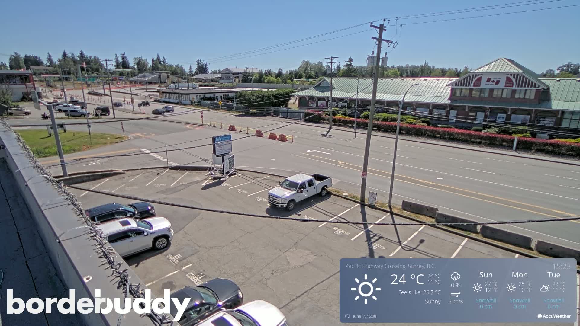 A sunny overhead view of a parking lot and roadway, with several vehicles parked and a few driving by, in front of some buildings.