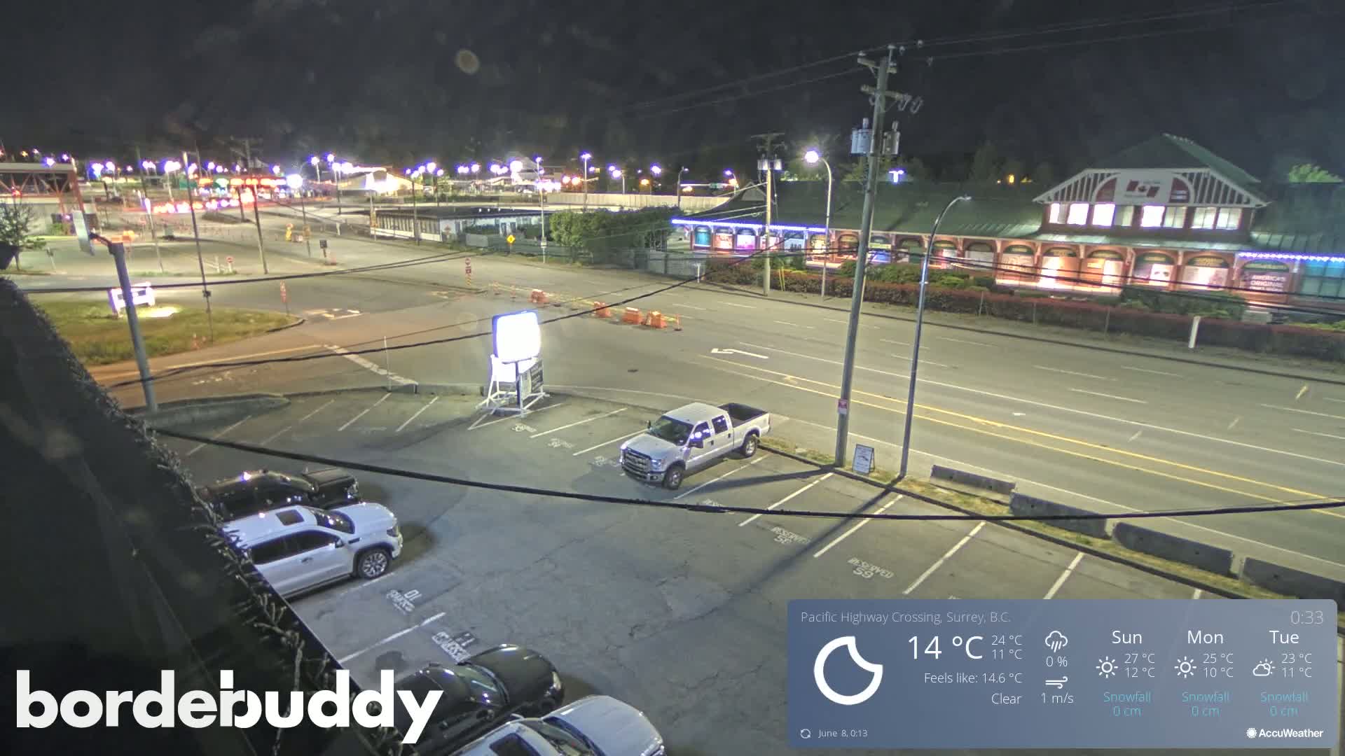 A nighttime, clear-weather view of a highway crossing, featuring several parked cars in a lot and a single pickup truck, with a building and some road construction visible in the distance.