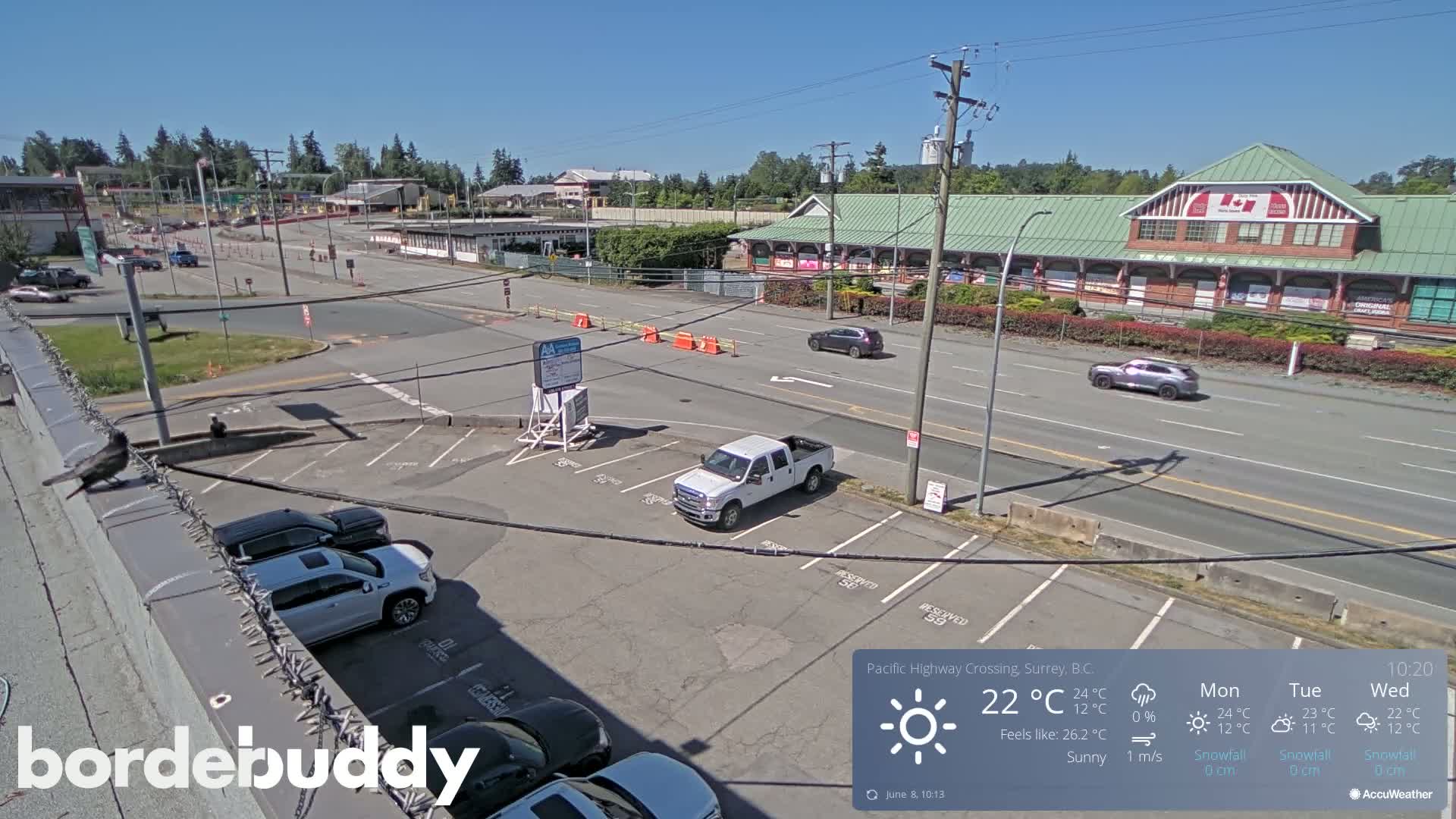 A sunny day is shown in a high-angle, long shot of a highway crossing with a parking lot and several vehicles, including cars and trucks.