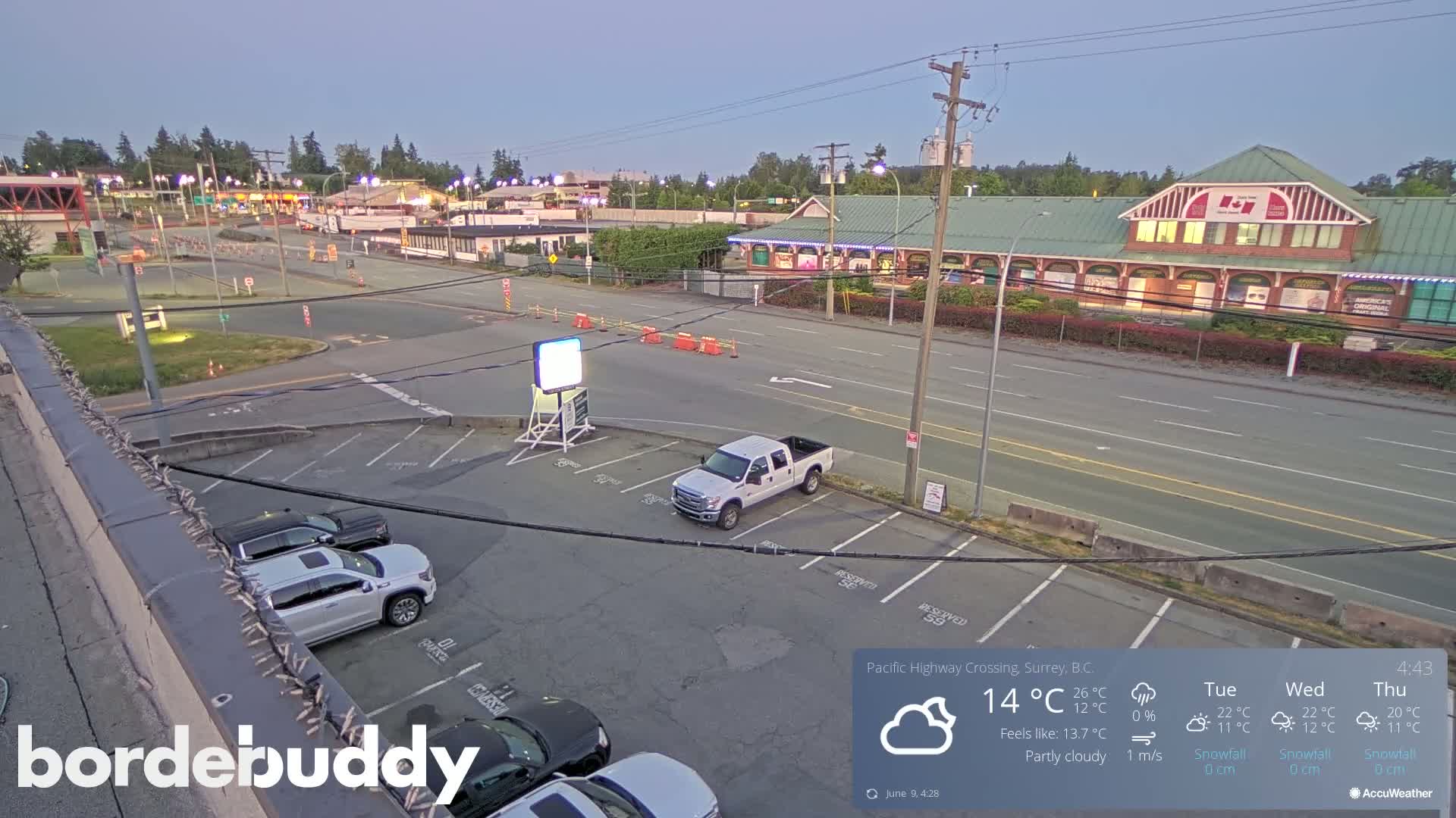 A partly cloudy evening scene shows a parking lot with several vehicles, a street with light traffic, and buildings in the distance.