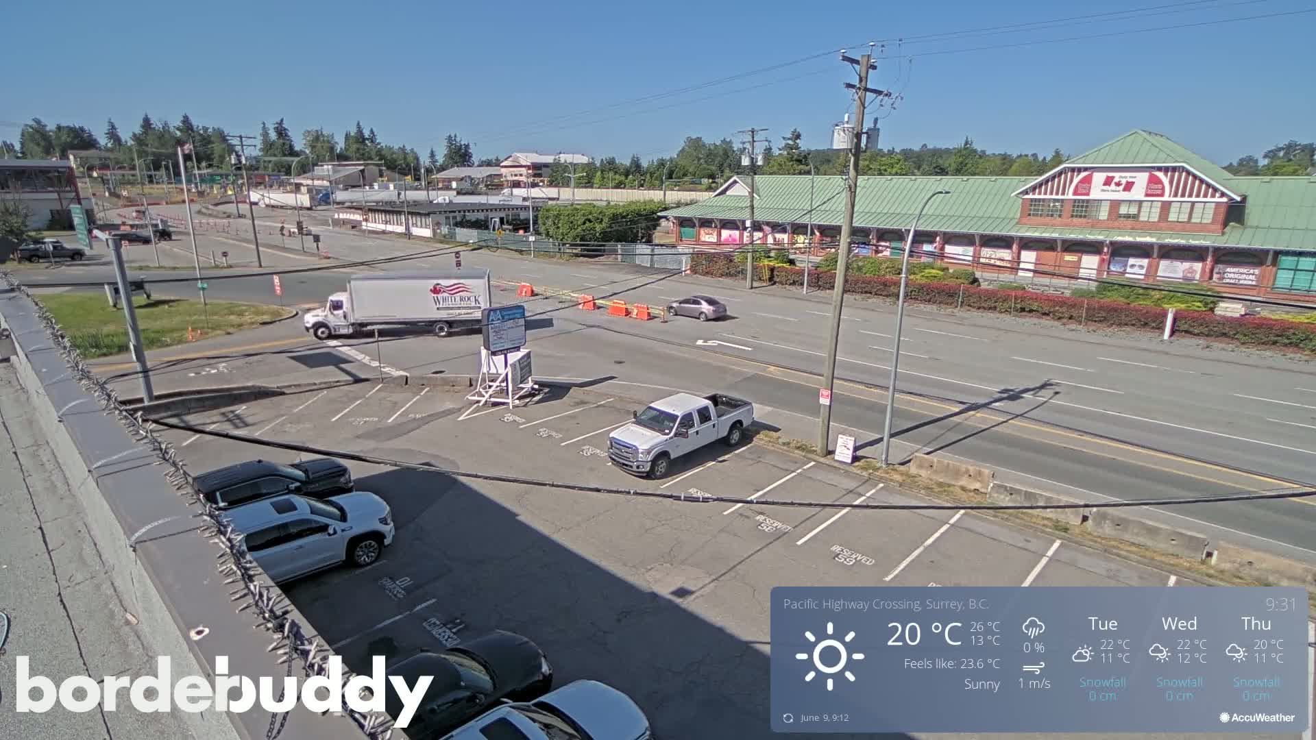 A sunny parking lot with several vehicles and a road leading to a large building complex under a clear sky.
