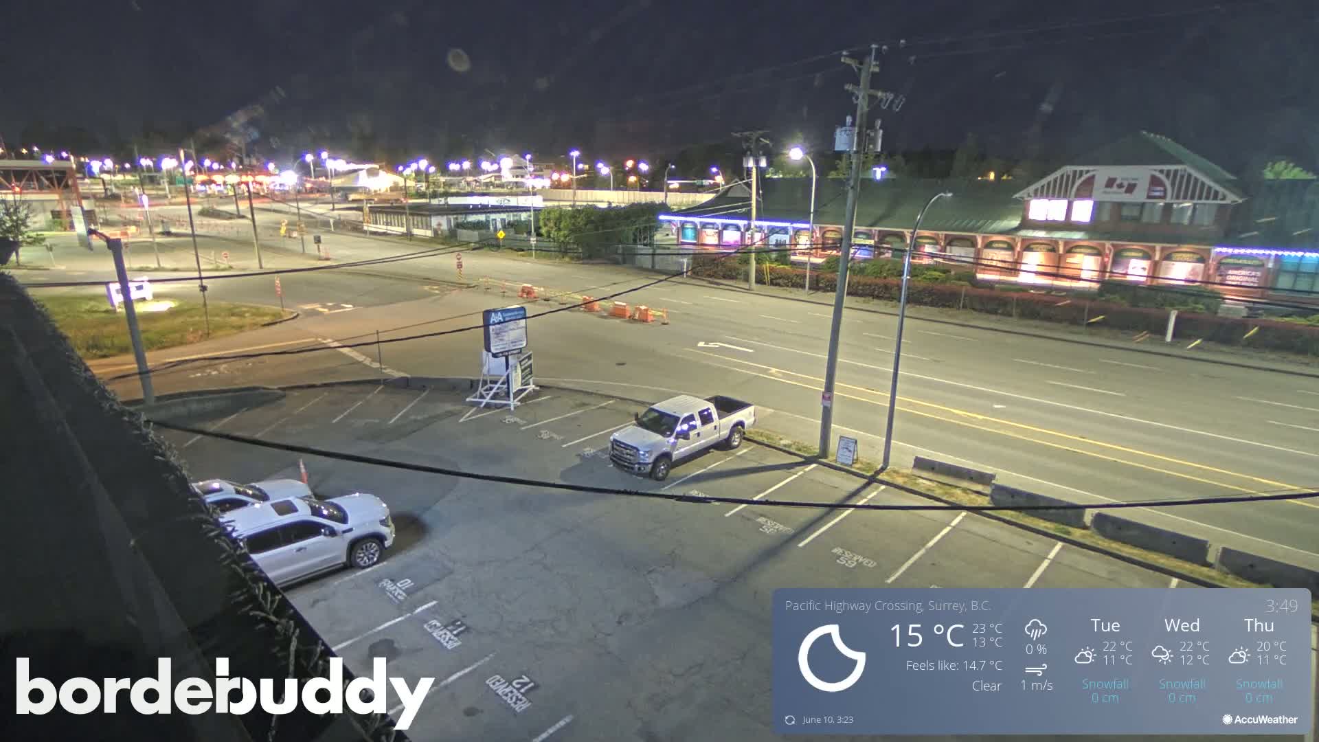 A nighttime, clear-weather view of a mostly empty parking lot, a roadway, and a building complex with some cars visible.