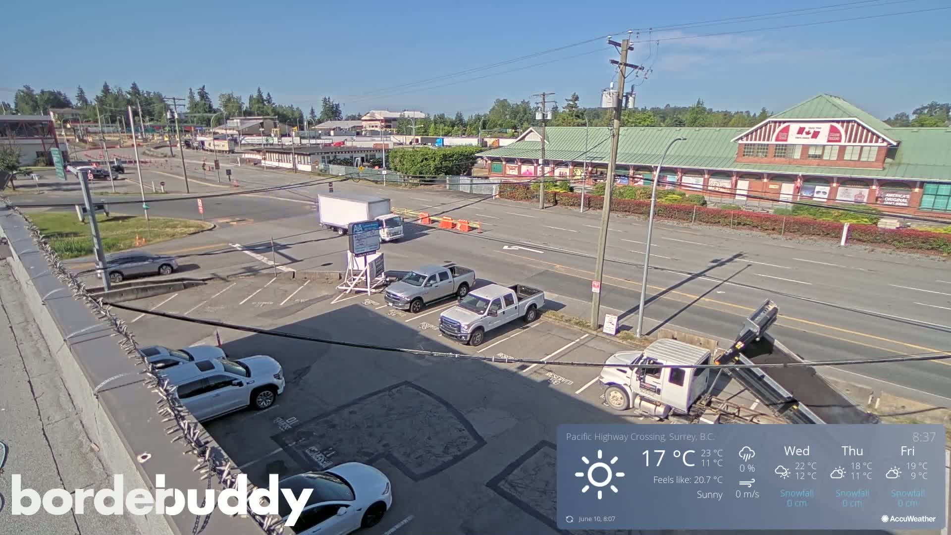 A sunny, overhead view of a paved road with several vehicles parked in a lot and others driving, in front of a long brick building.
