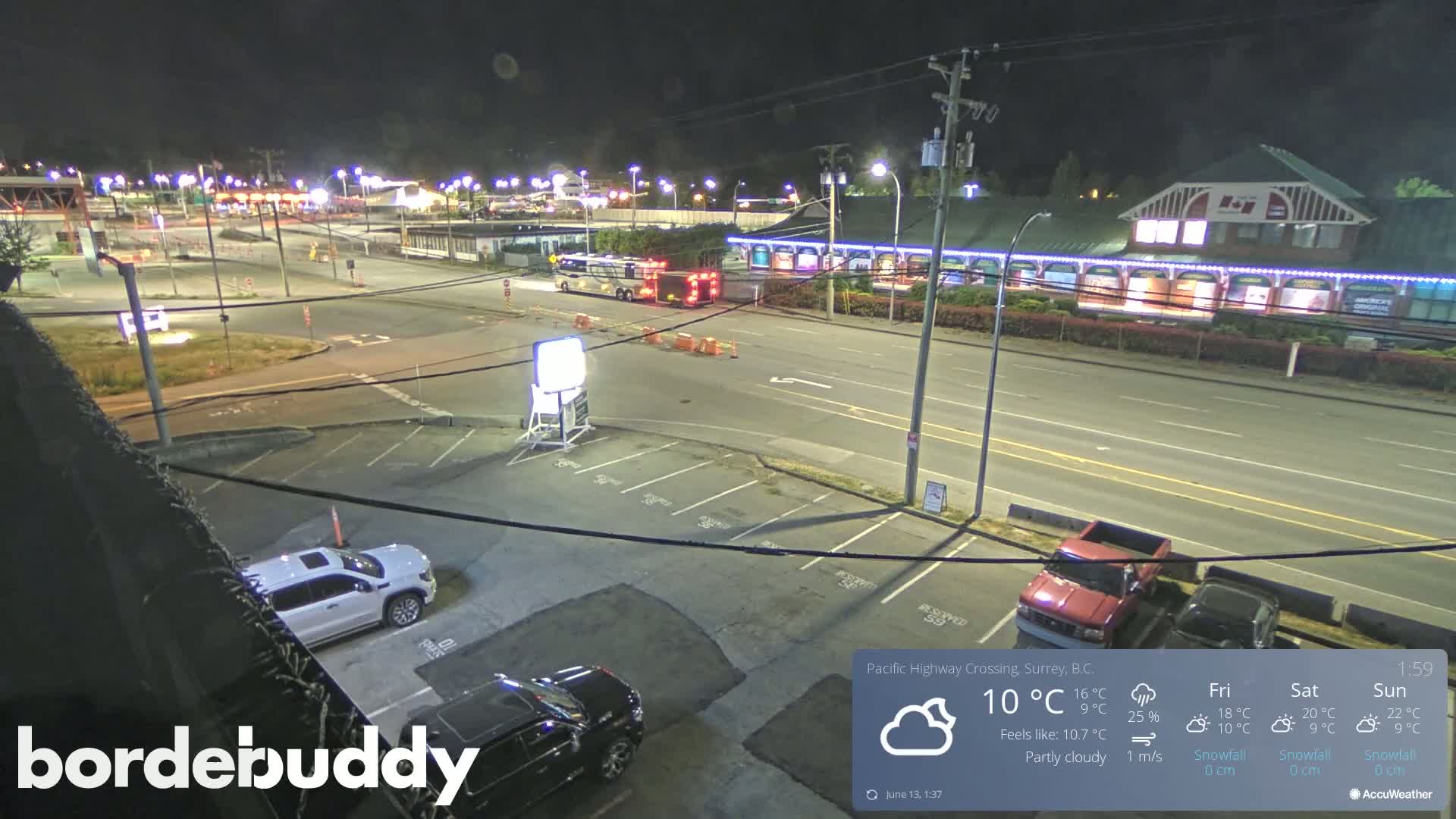 A nighttime, partly cloudy scene shows a roadway intersection with parked cars in a lot, a building with lights, and a large vehicle near a traffic signal.