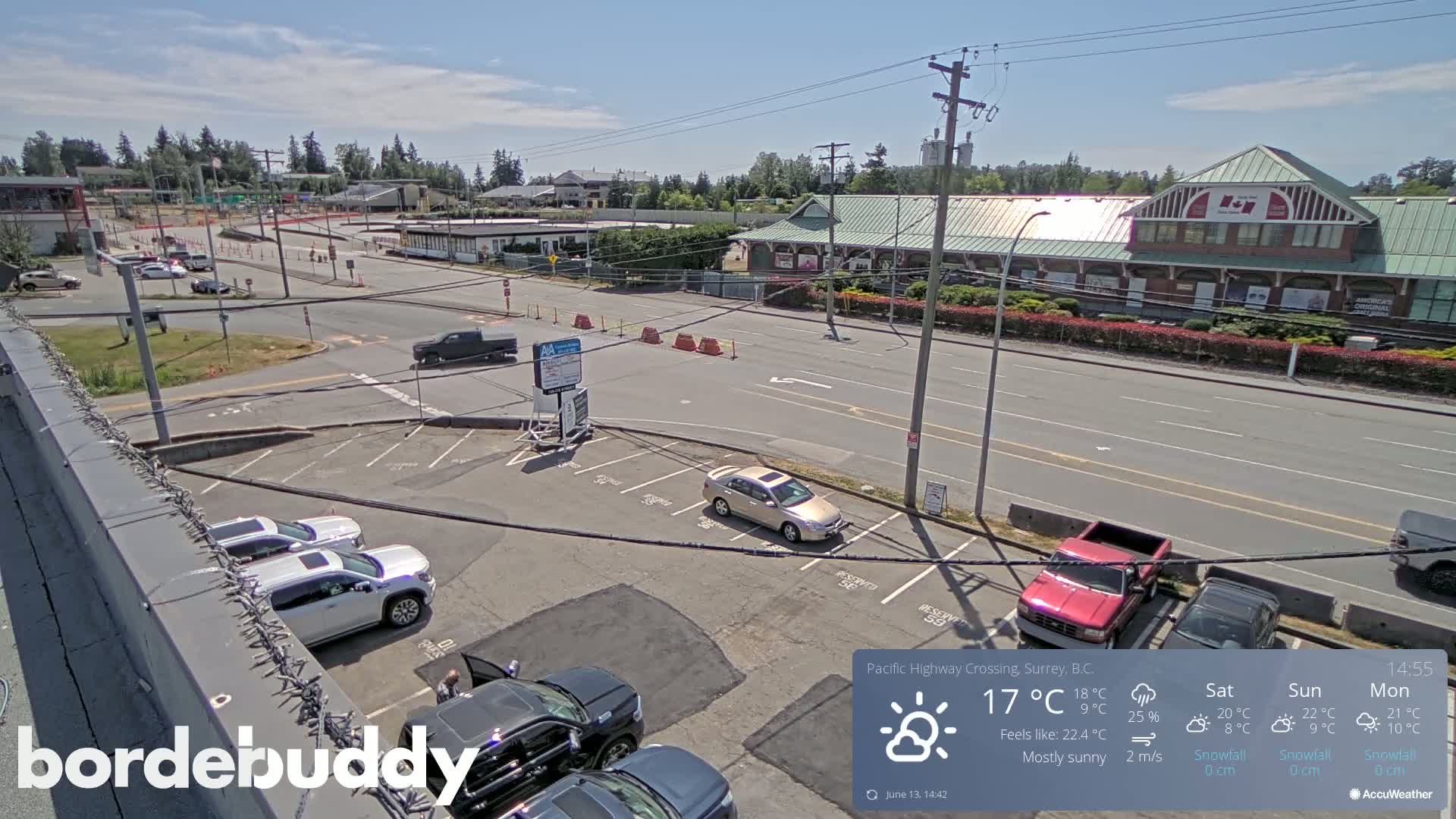 A mostly sunny parking lot and street scene shows several cars parked in a lot adjacent to a busy roadway, with some buildings in the background.