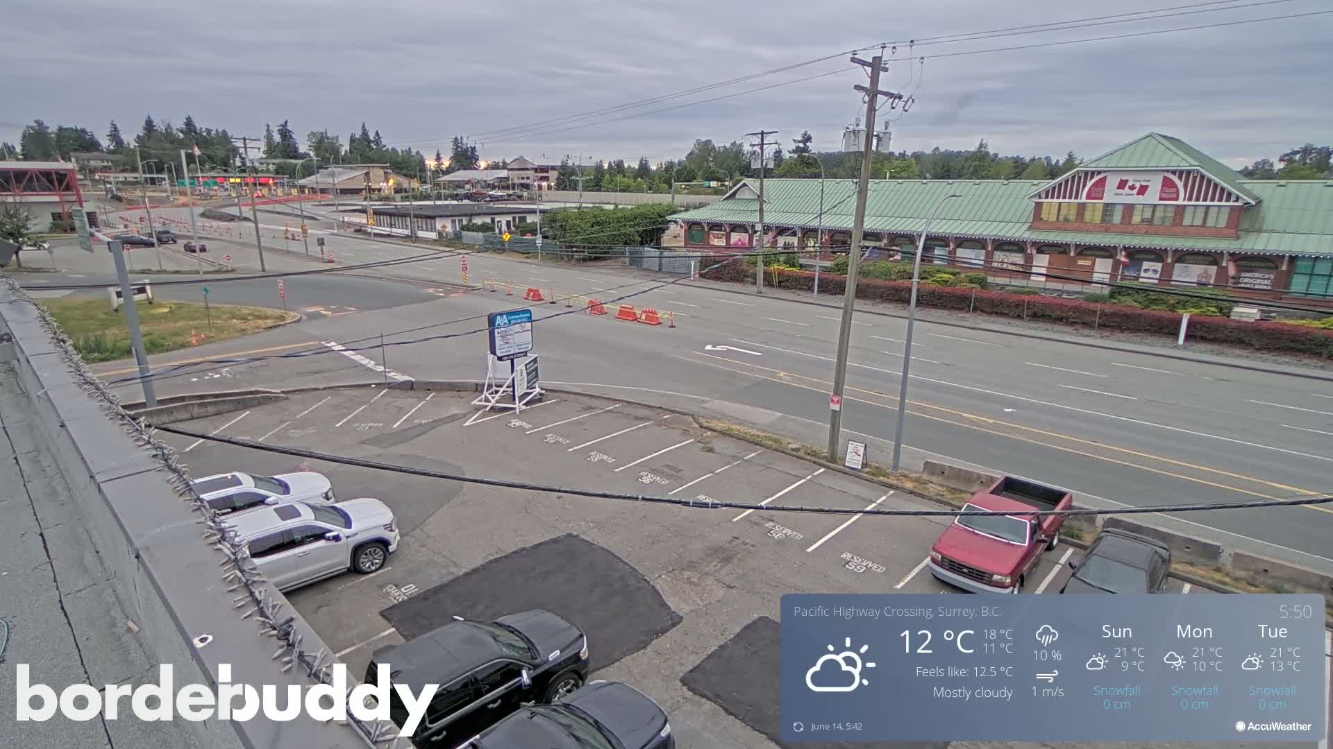 A mostly cloudy daytime scene shows a parking lot with several vehicles, a roadway with light traffic, and buildings in the background.