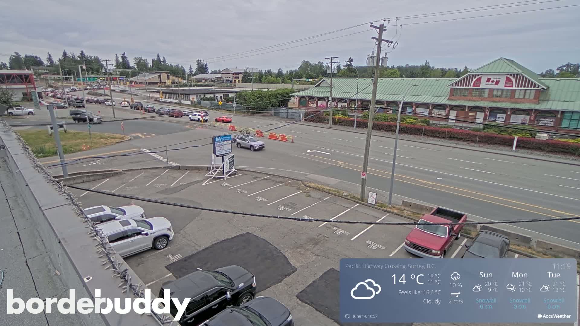 A cloudy day shows a street scene with cars parked in a lot and driving on a road in front of buildings, one of which is a large market.