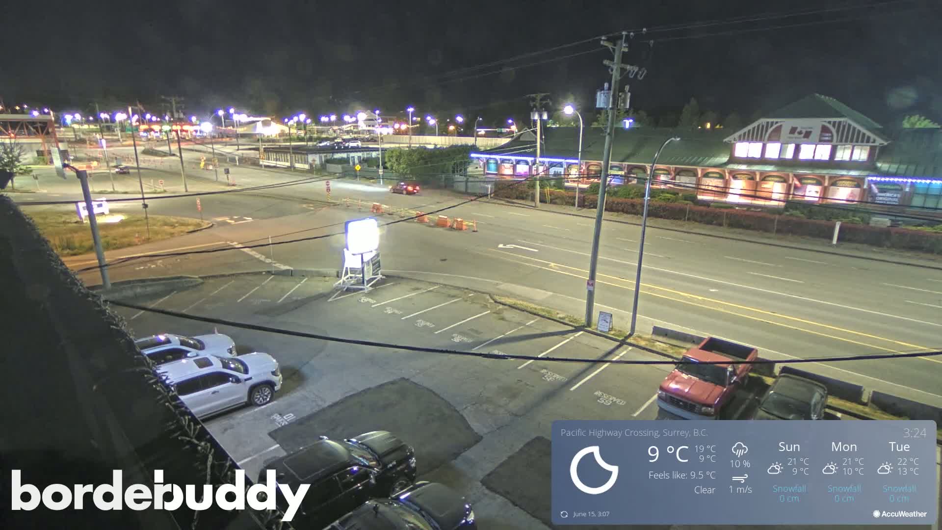 A nighttime, clear-weather view of a parking lot and roadway, showing several vehicles and buildings under streetlights.