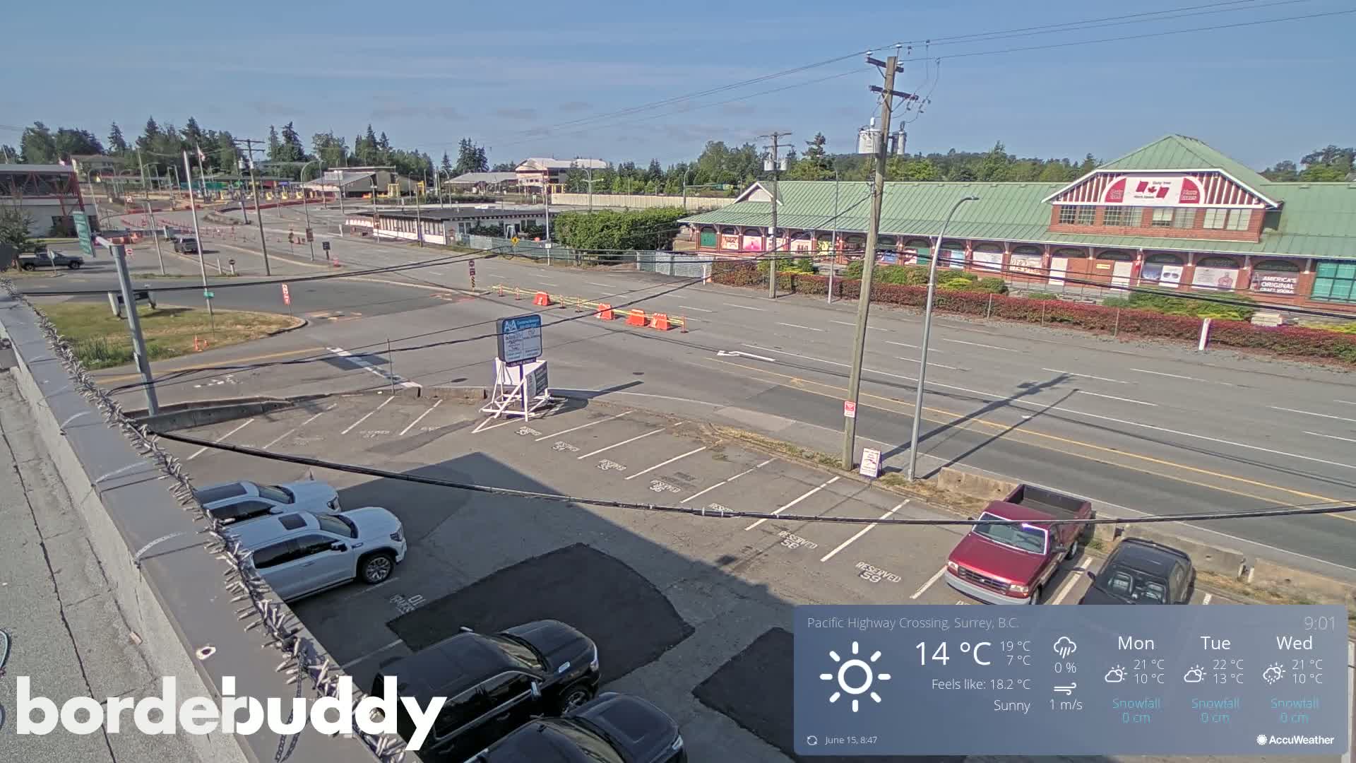 A sunny, overhead view of a highway crossing with several cars parked in a lot and buildings in the distance.