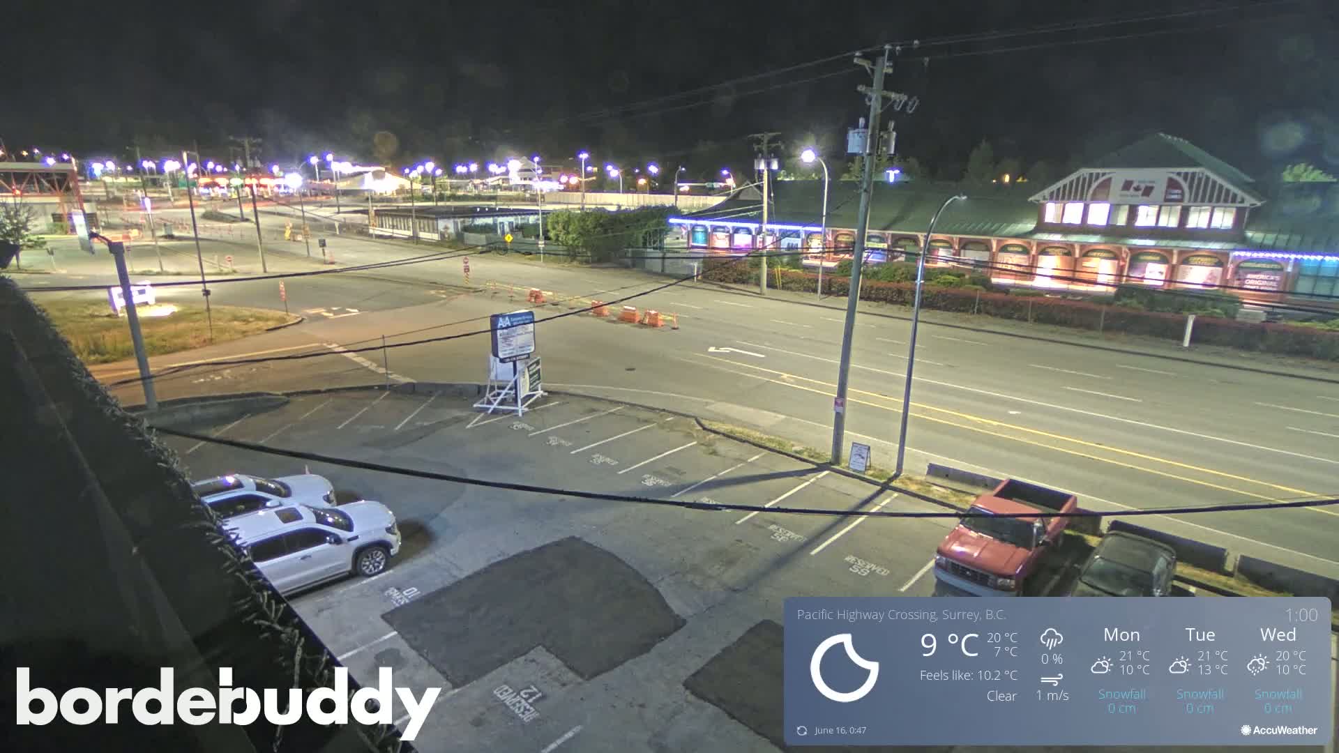 A nighttime, clear view of a mostly empty parking lot and street, with several vehicles parked, and buildings and streetlights visible in the distance.