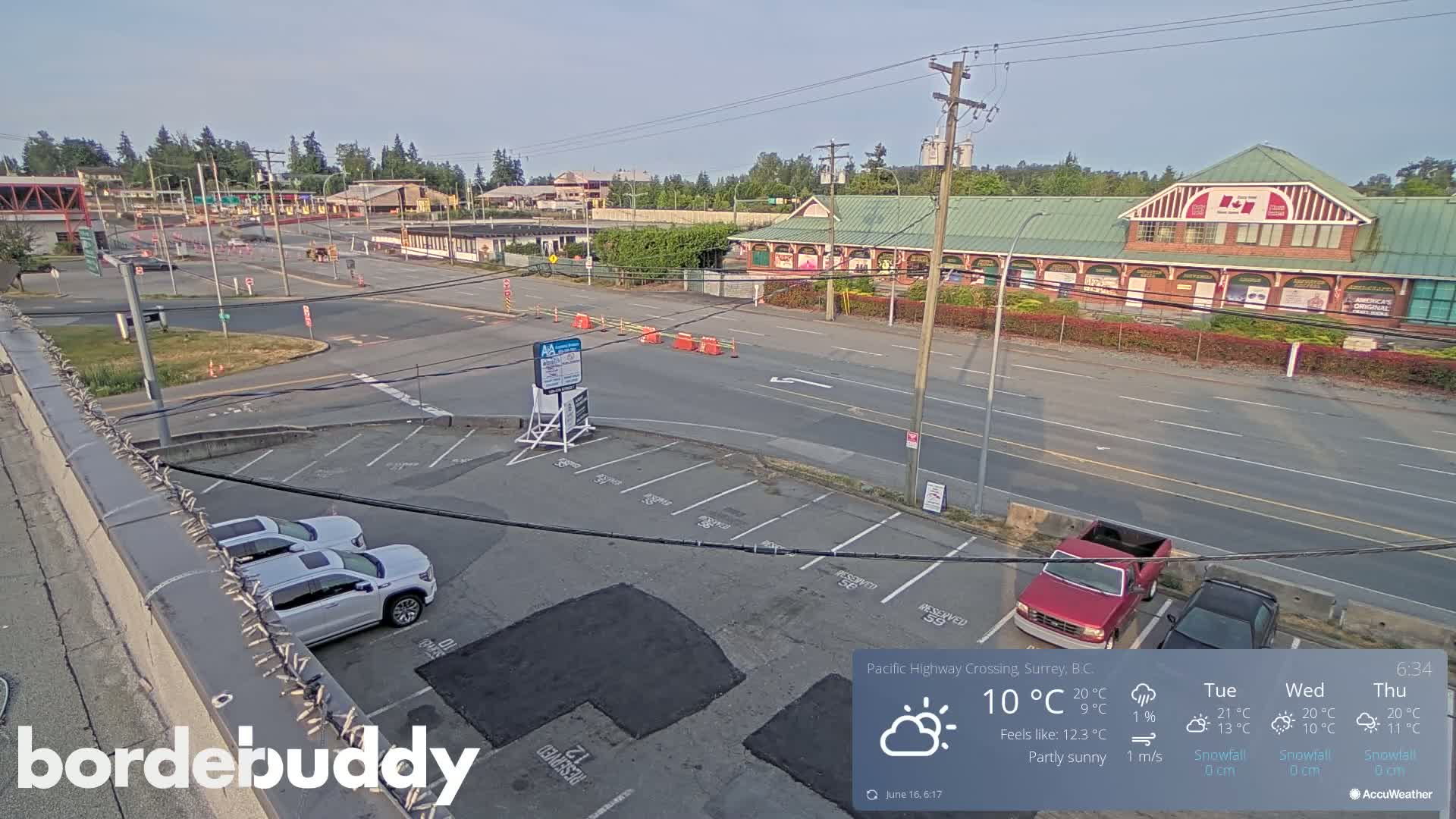 A partly sunny view of a roadway and parking lot with several vehicles, a building with a green roof, and road construction visible in the distance.