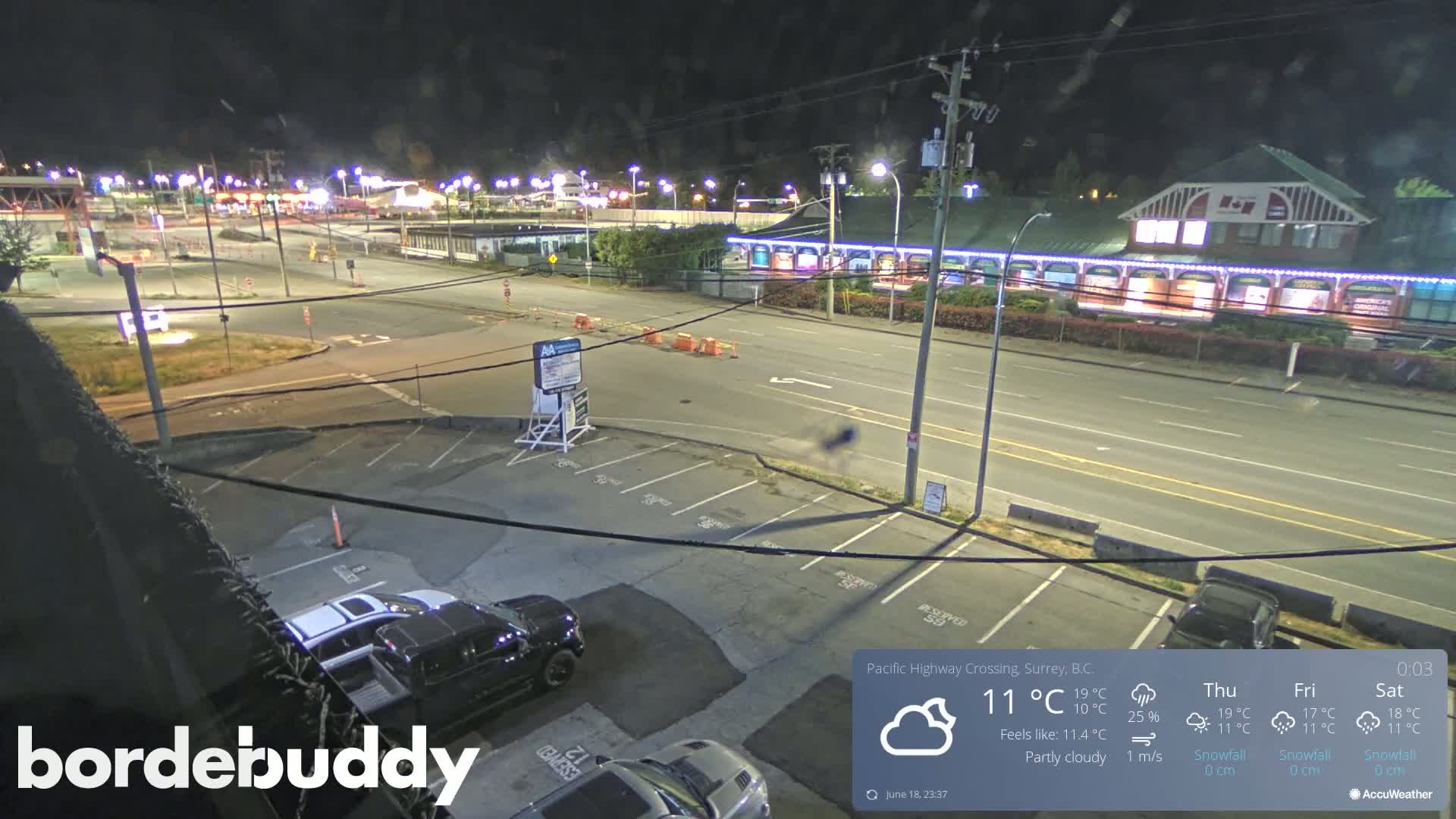 A nighttime, partly cloudy view of a parking lot and roadway next to a building, with several vehicles visible.