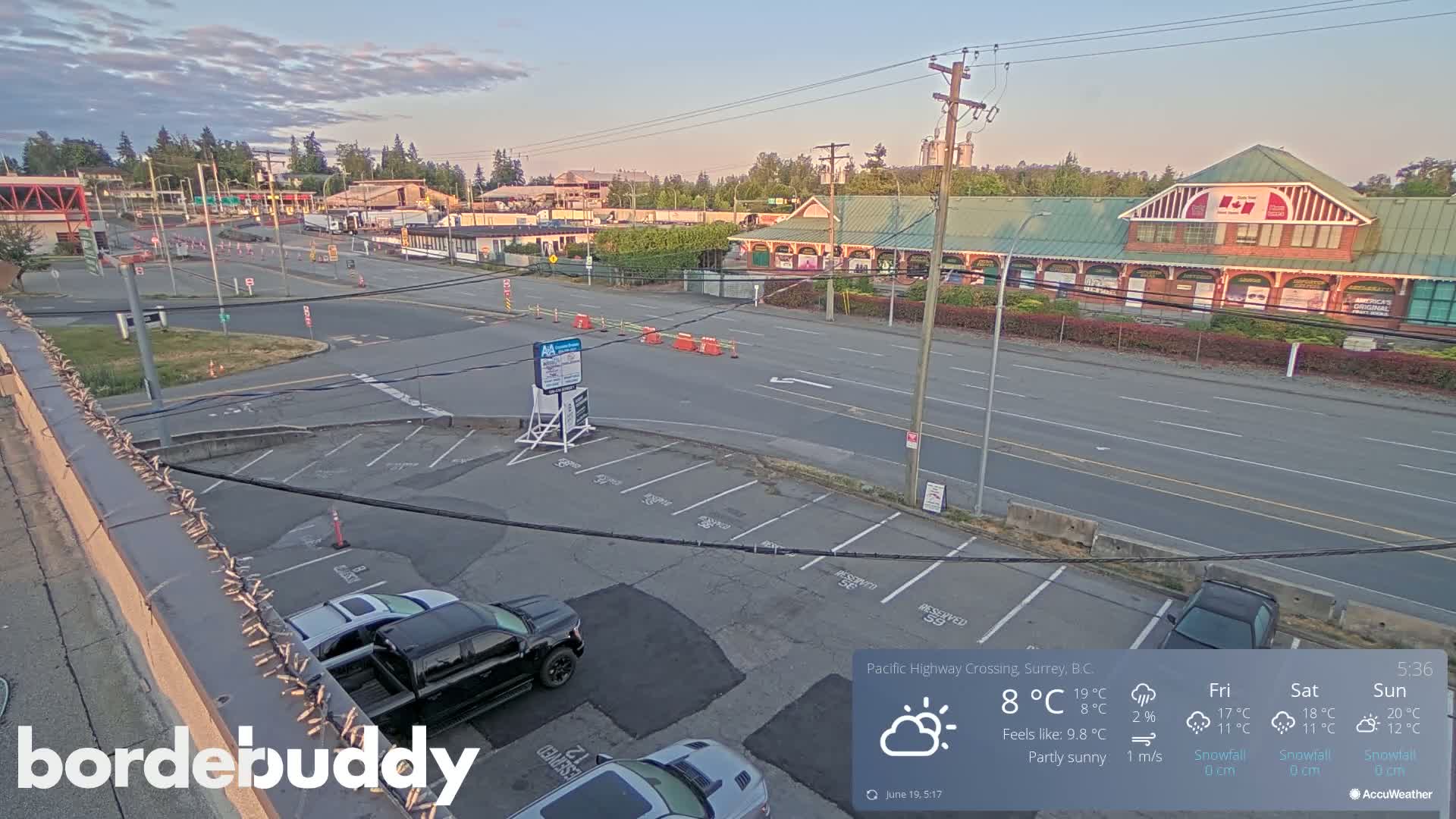A partly sunny view of a highway crossing with a parking lot and several buildings, including a long brick building with a green roof.