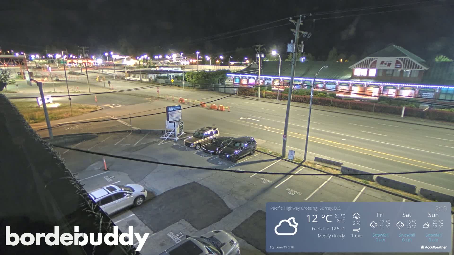 A mostly cloudy night scene shows a parking lot with several cars, and a road leading to a brightly lit building complex.