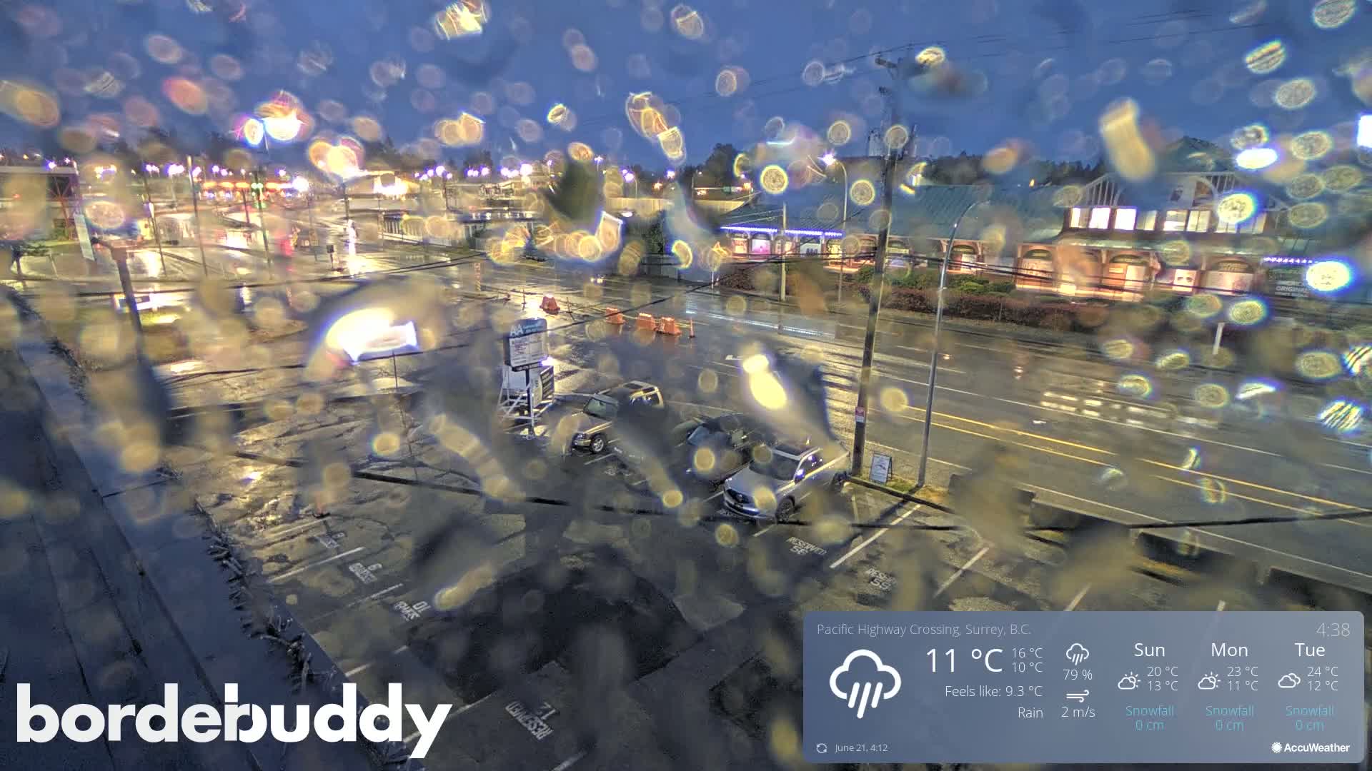 Rain falls on a wet parking lot at night, viewed through a rain-covered window, showing several cars parked and a building in the background.