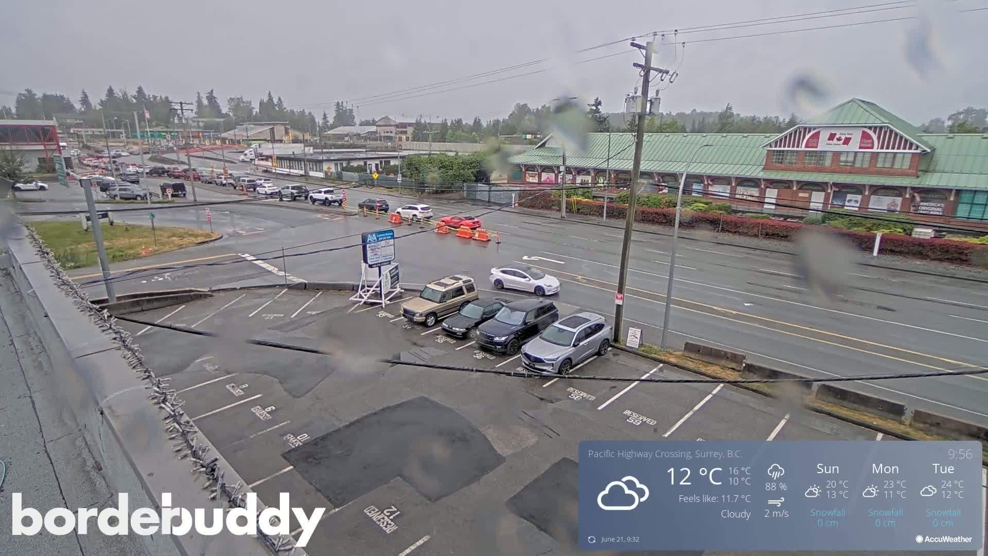 A wet parking lot with several cars is seen next to a roadway with other vehicles and buildings under a cloudy sky.