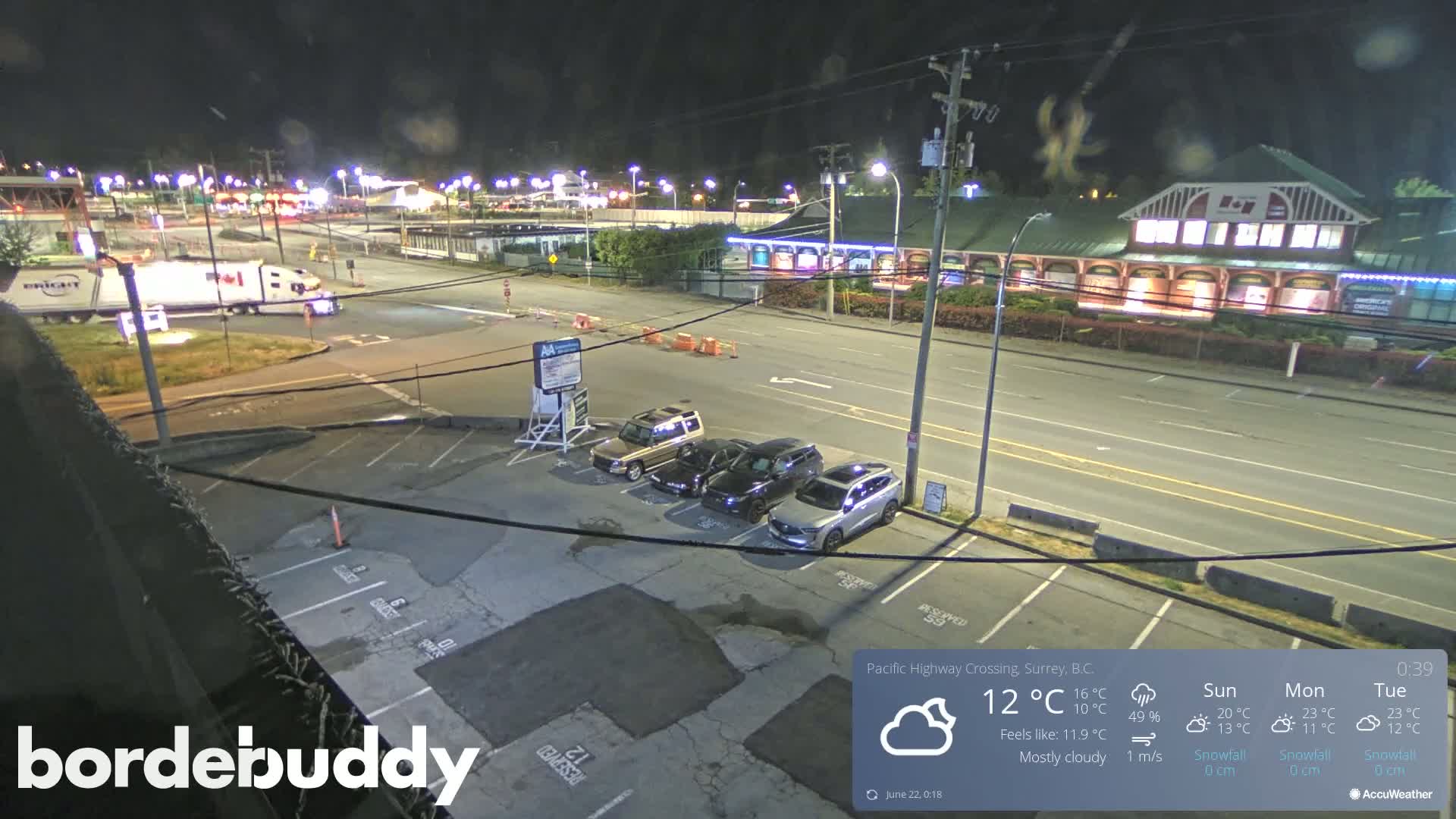 A mostly cloudy night scene shows a parking lot with several cars, a roadway with a large truck, and a building in the background.